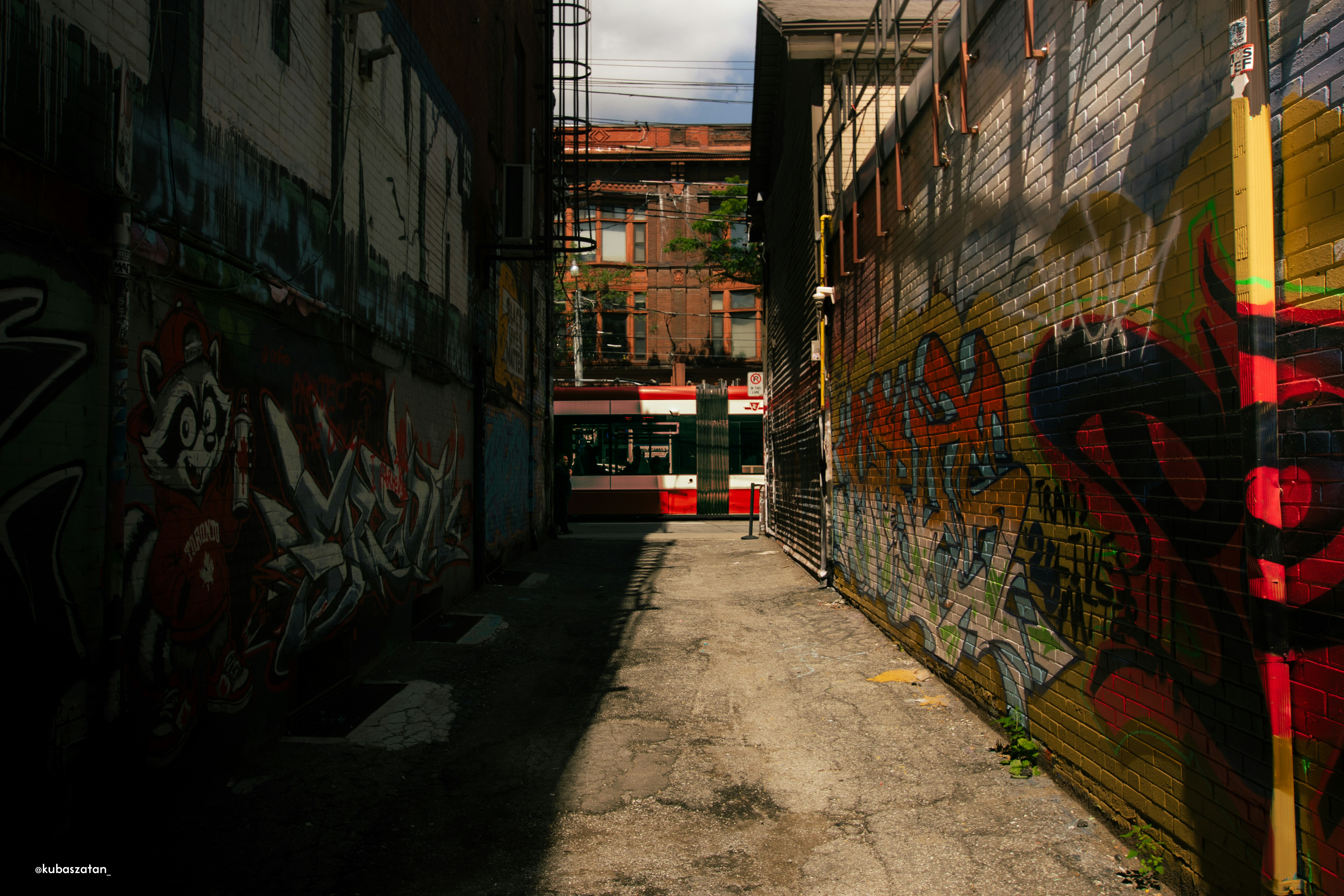 A narrow alleyway with graffiti-covered brick walls and a red streetcar passing in the distance