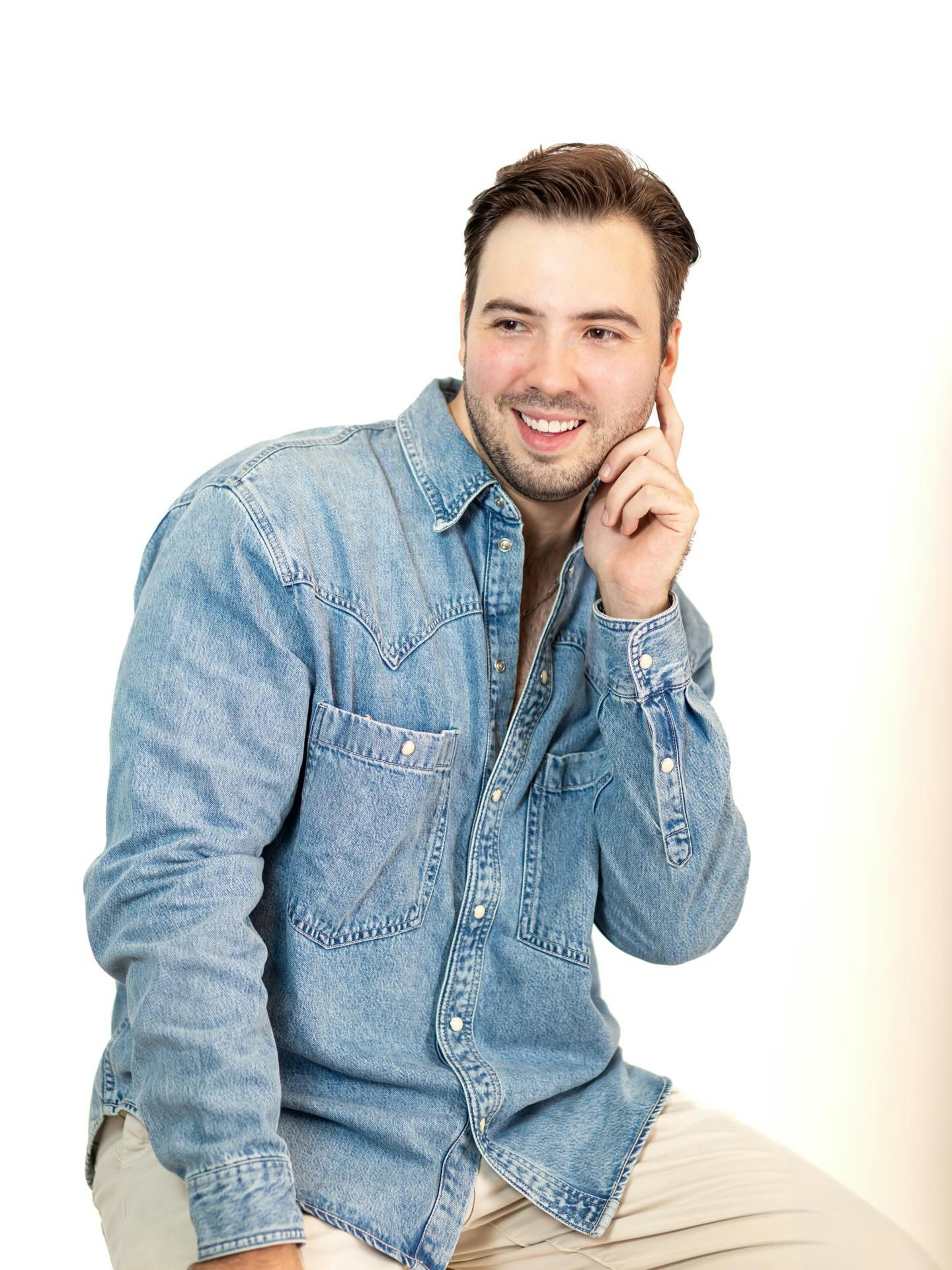 A smiling man in a denim shirt with his hand to his face