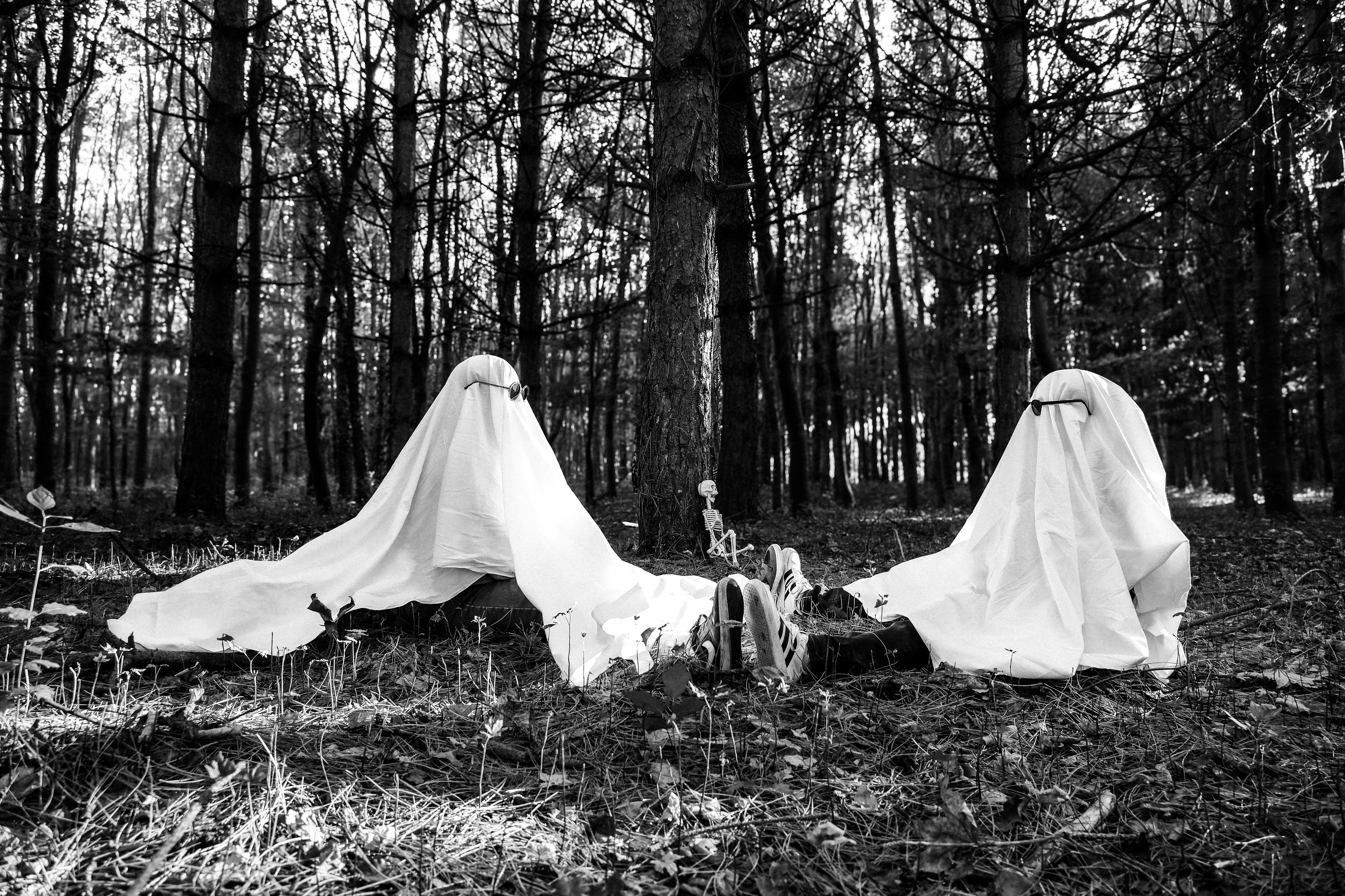 Two people in ghost costumes with sunglasses sitting on the forest floor near a small skeleton model