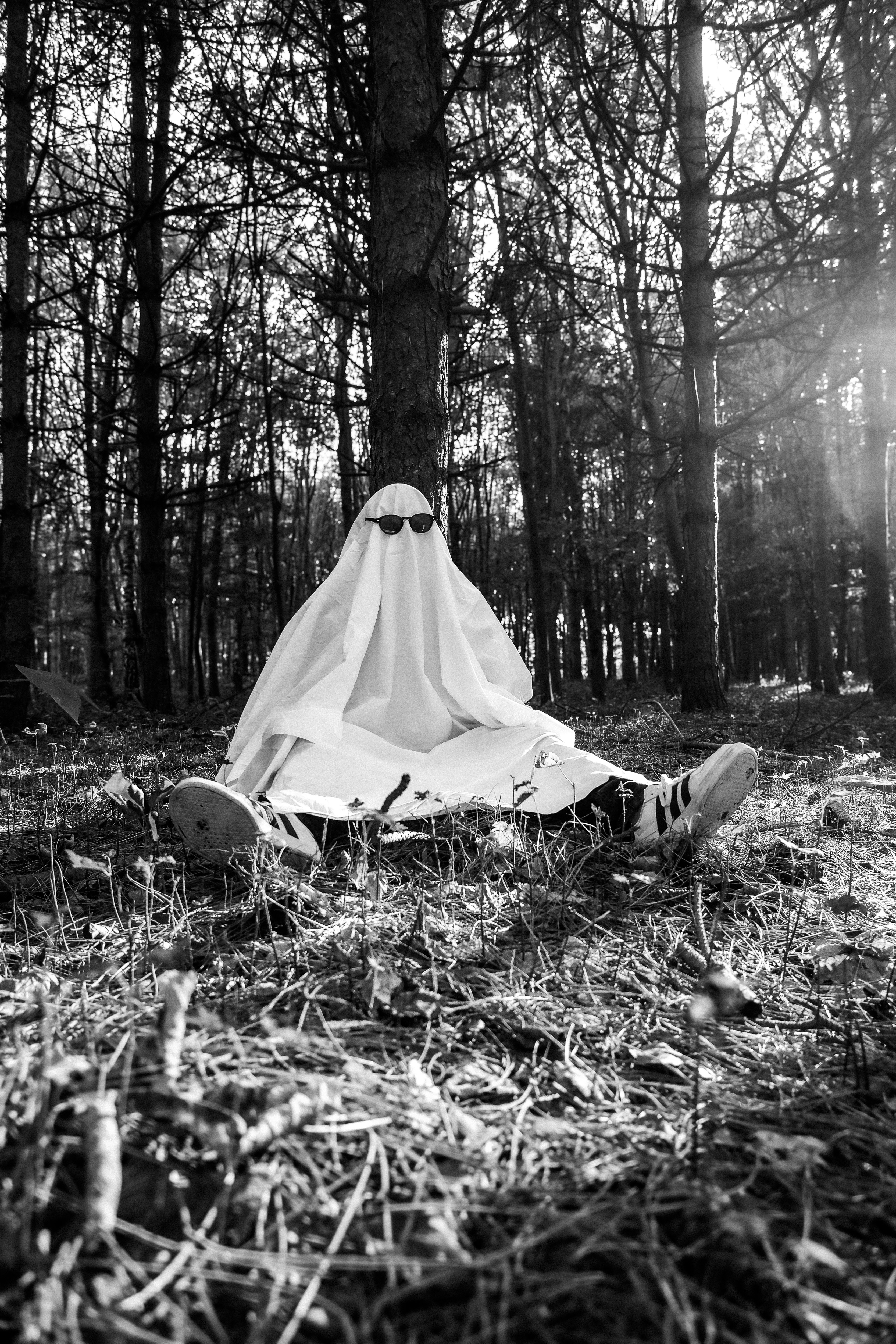 A person in a white sheet ghost costume and sunglasses sitting against a tree in a forest