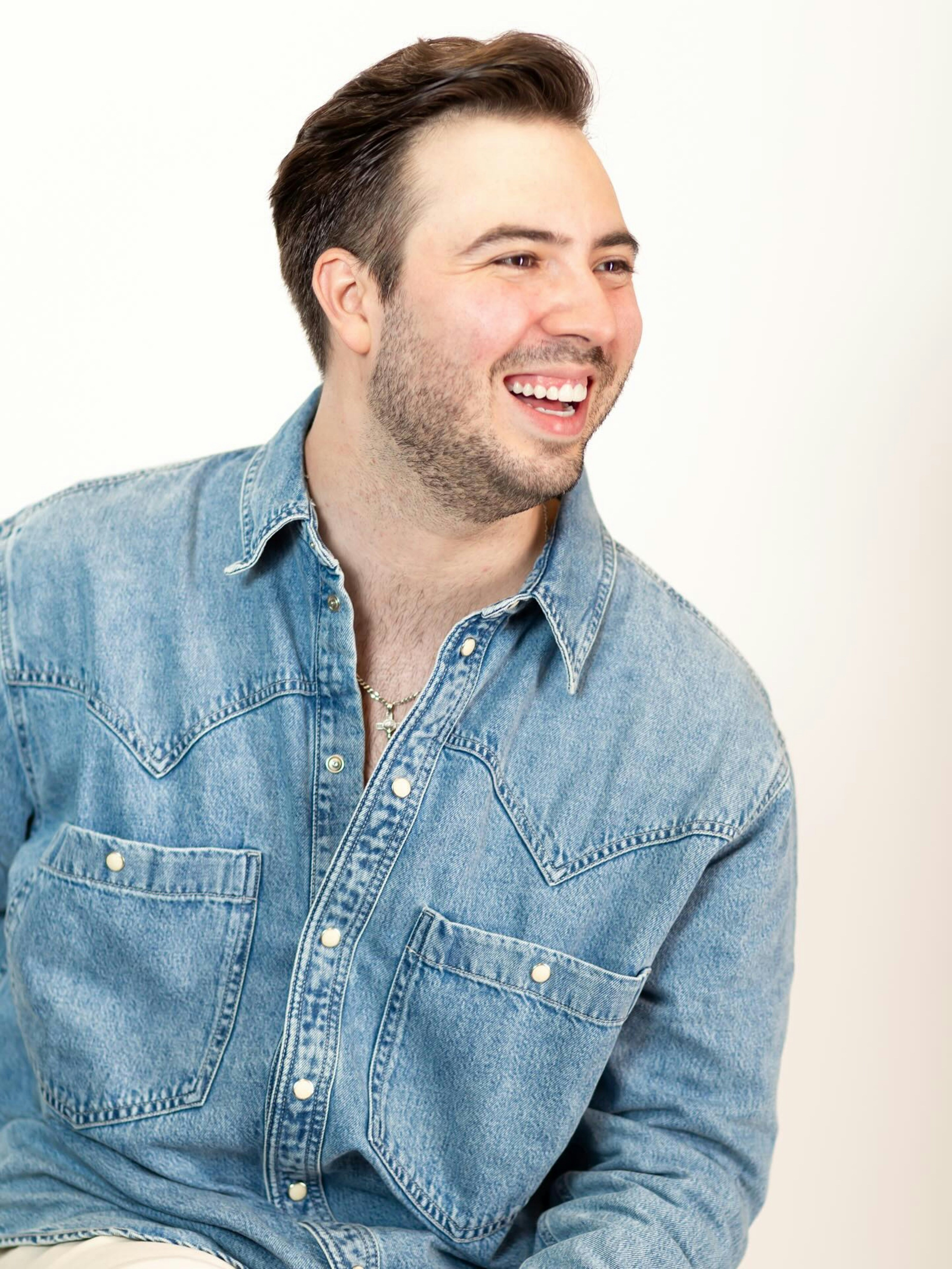 A laughing adult man with a beard wearing a blue denim shirt against a white background