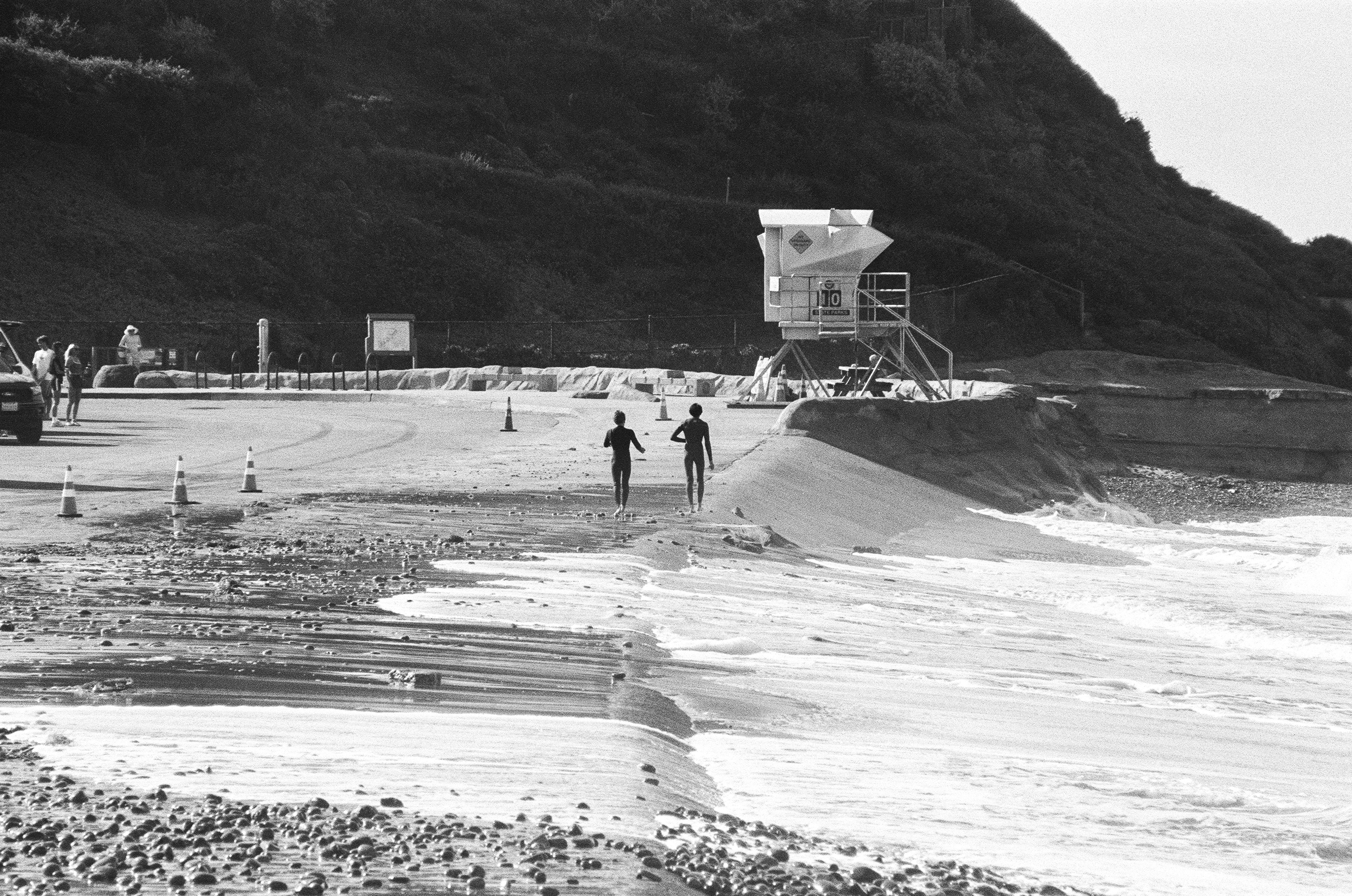 Two people in wetsuits walking along a beach past a lifeguard tower in black and white