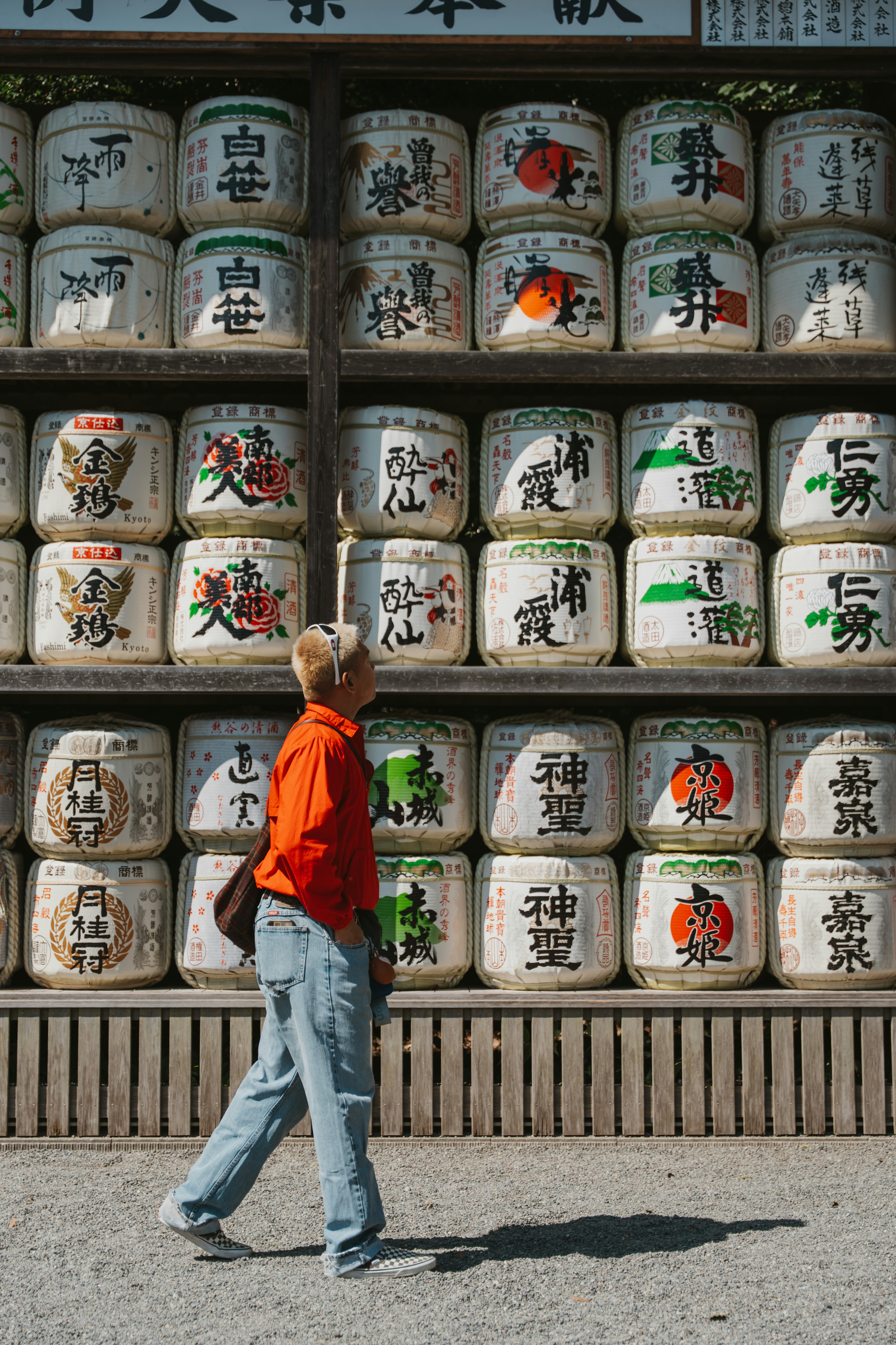 A person in an orange shirt walking past a wall of stacked sake barrels in Japan