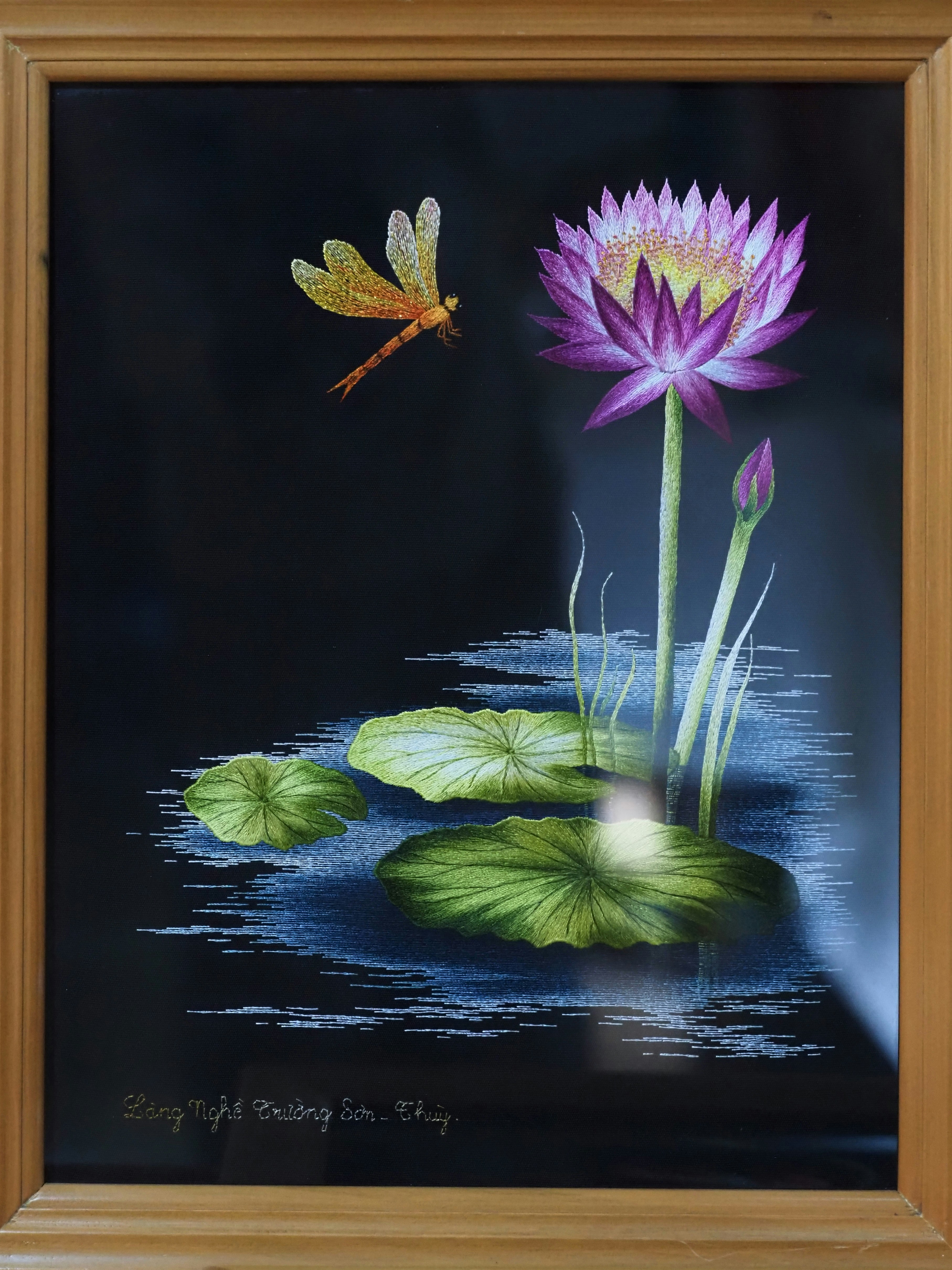 An embroidery of a purple lotus flower and a dragonfly over water on a black background
