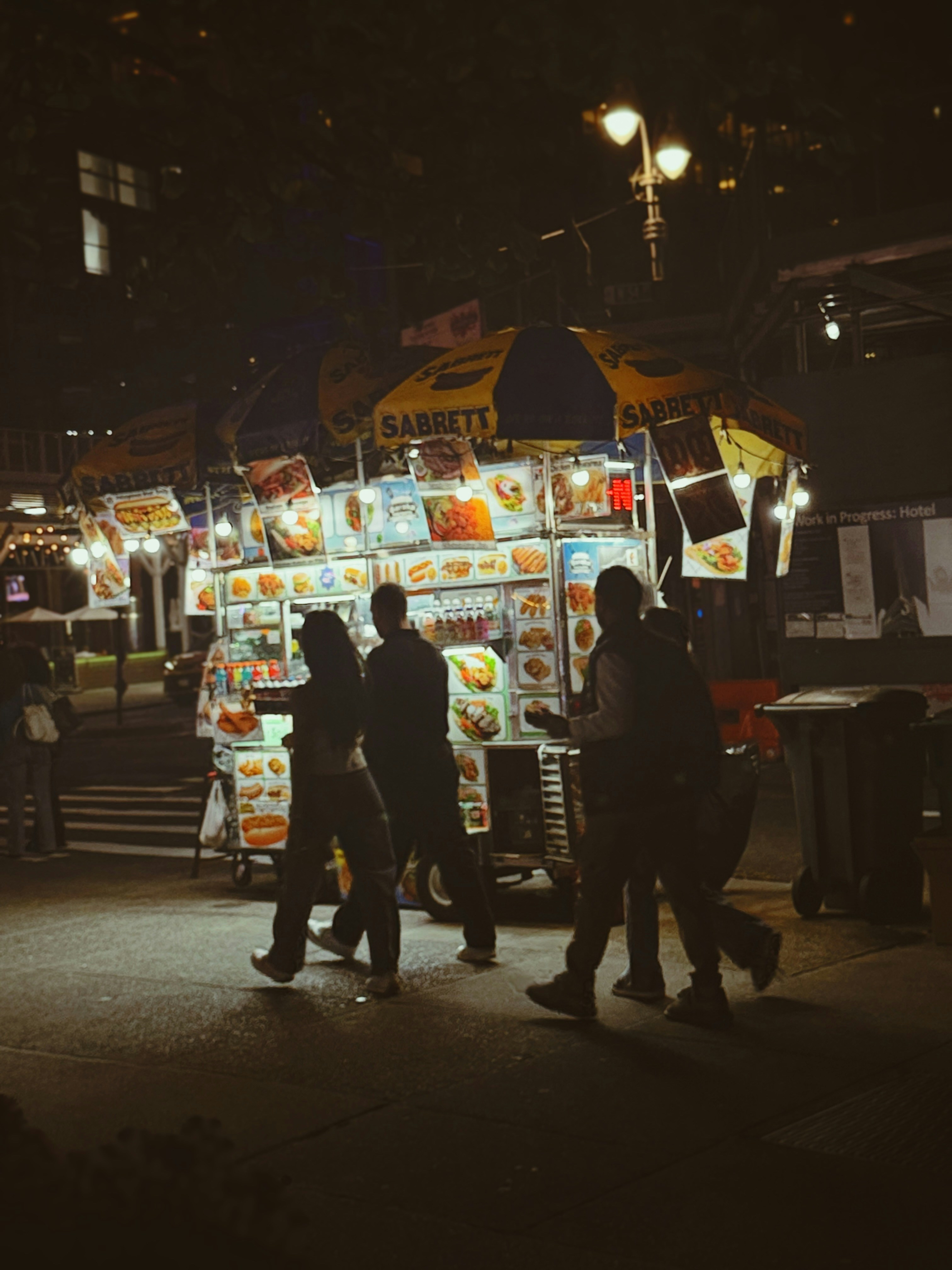 People walking past a brightly lit street food cart with a yellow umbrella at night
