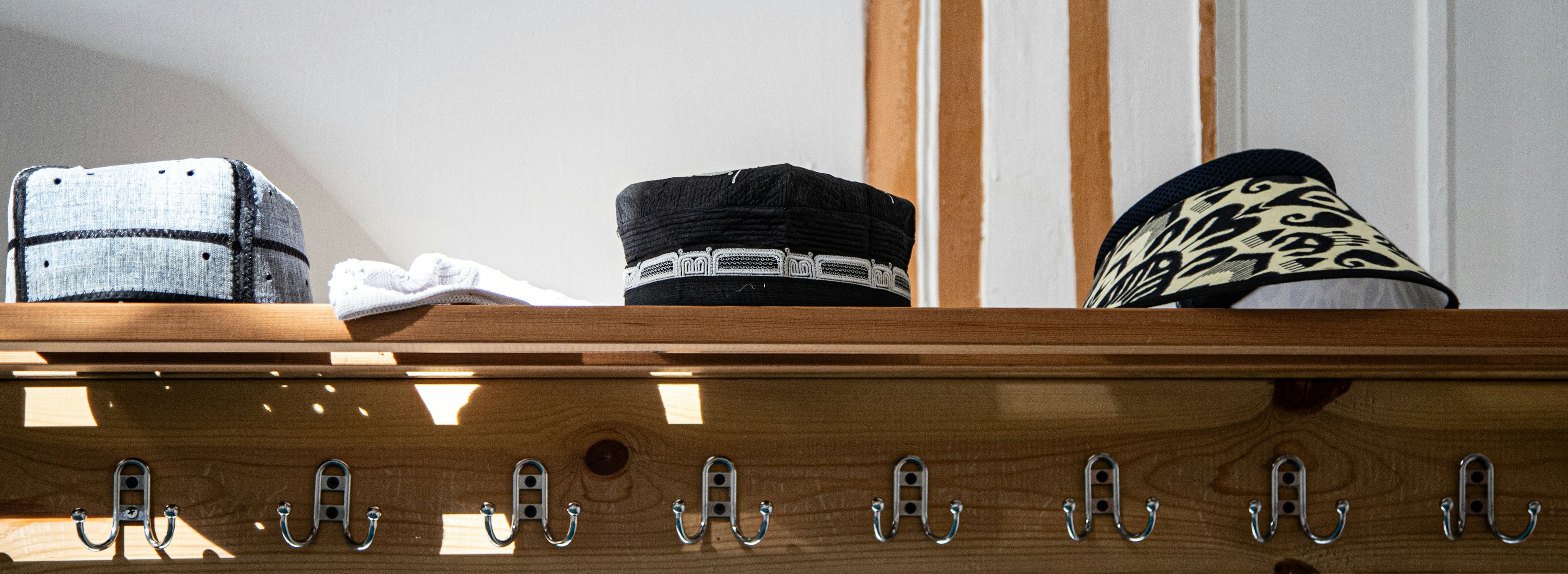 Three hats on a wooden shelf above a row of metal coat hooks