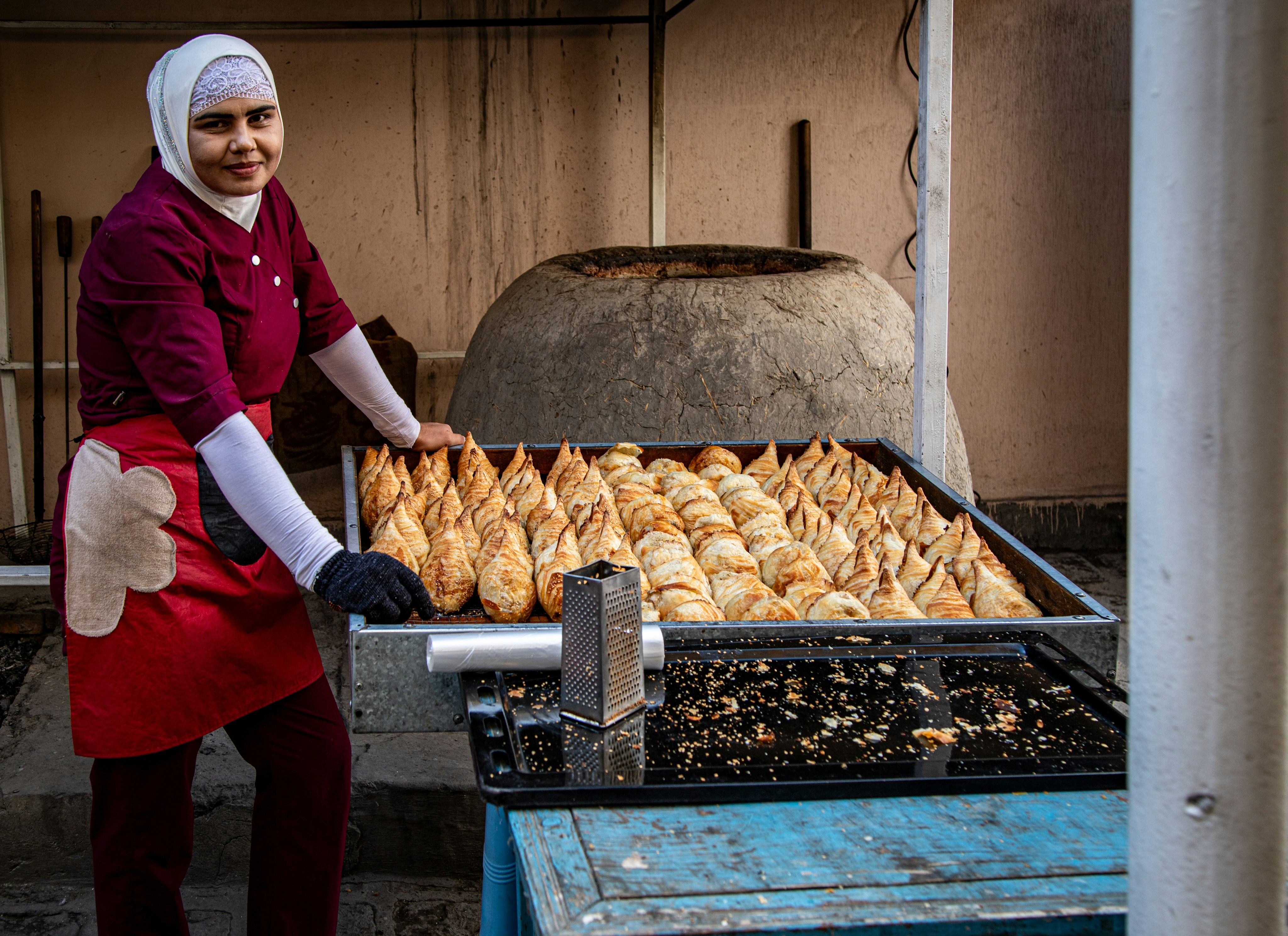 A woman in a hijab and apron standing beside a tray of baked pastries near a tandoor oven