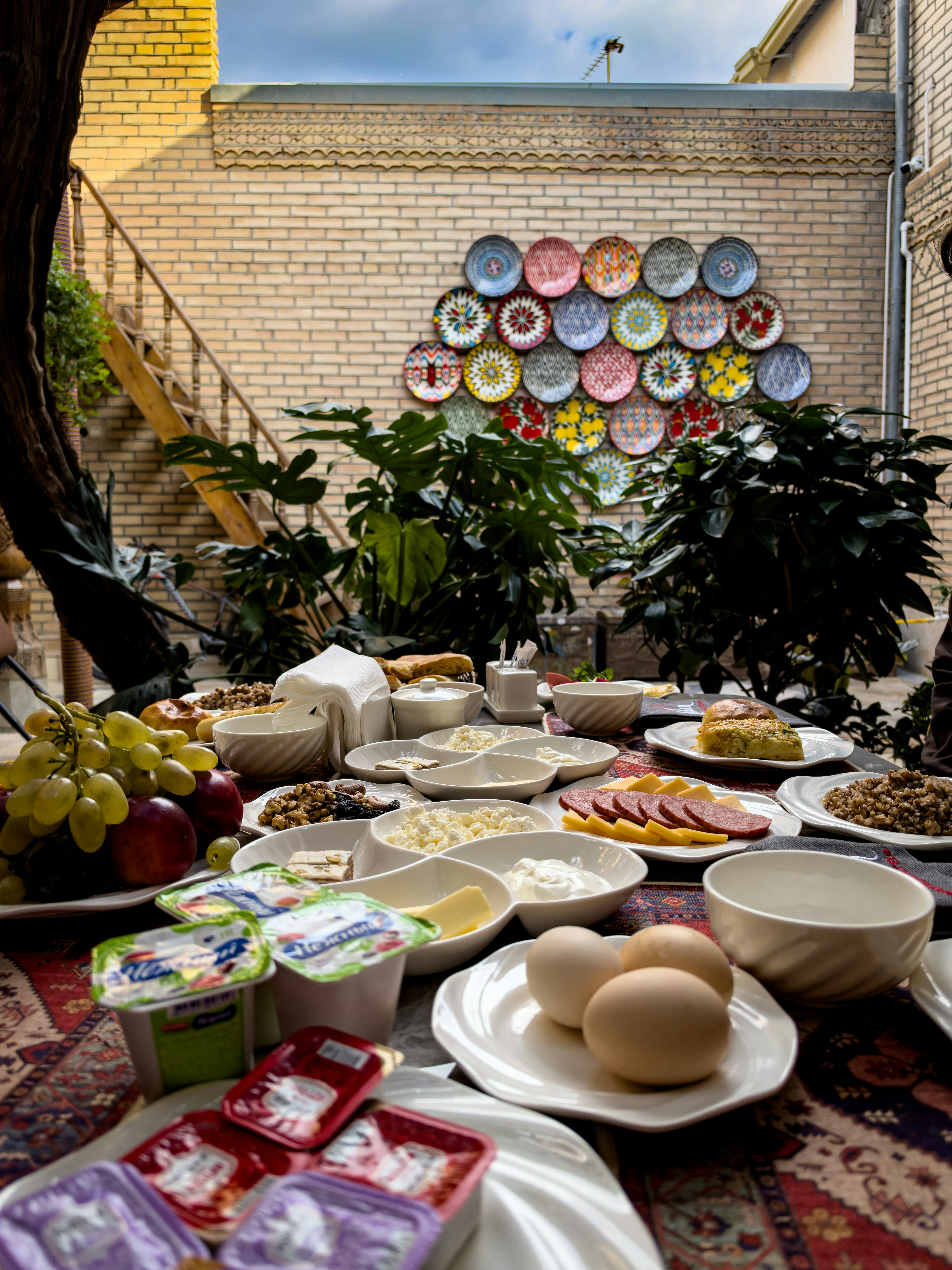A breakfast spread with eggs, cheese, fruit, and meats on a table in a courtyard
