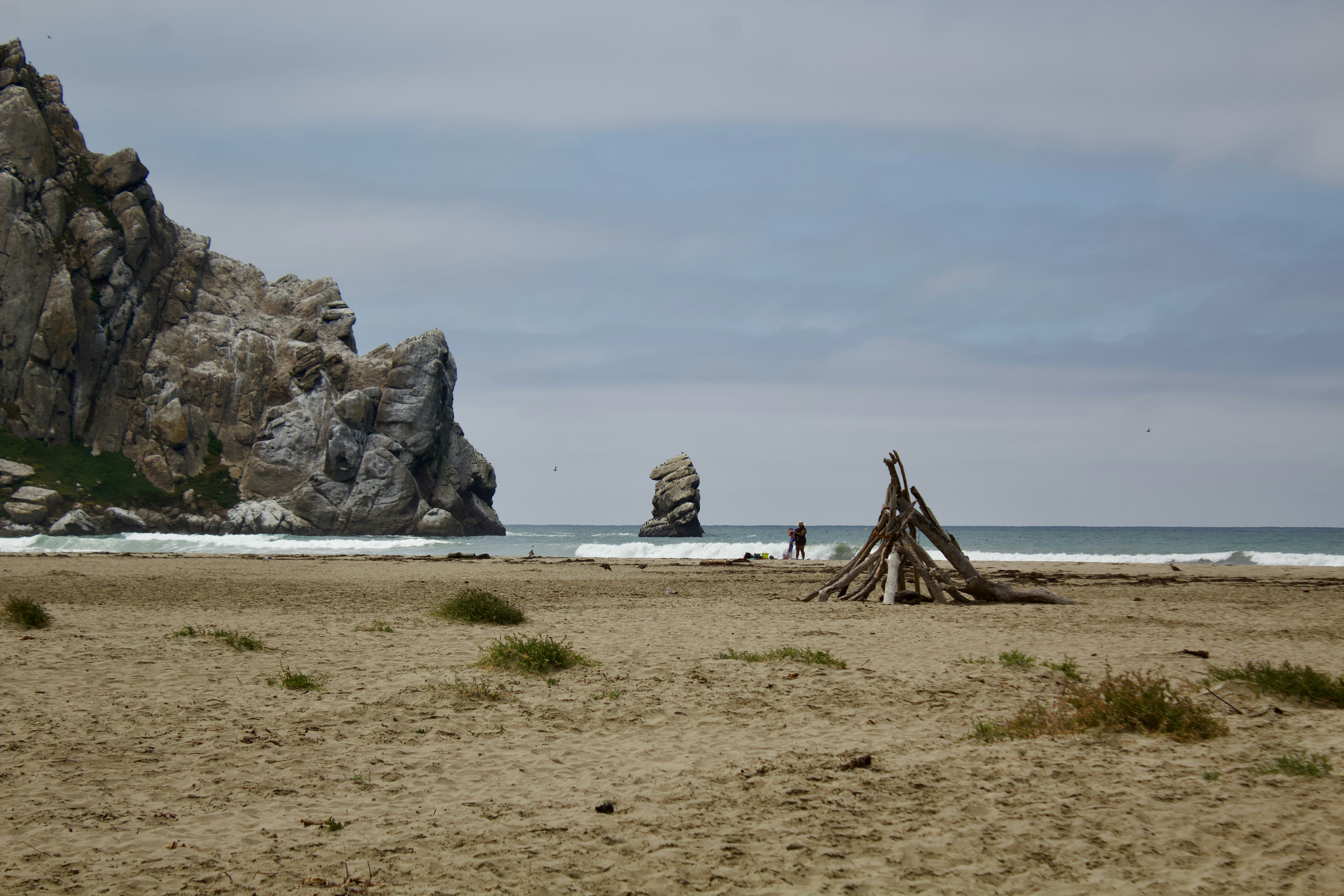A driftwood structure on a sandy beach with a large rock formation in the distance