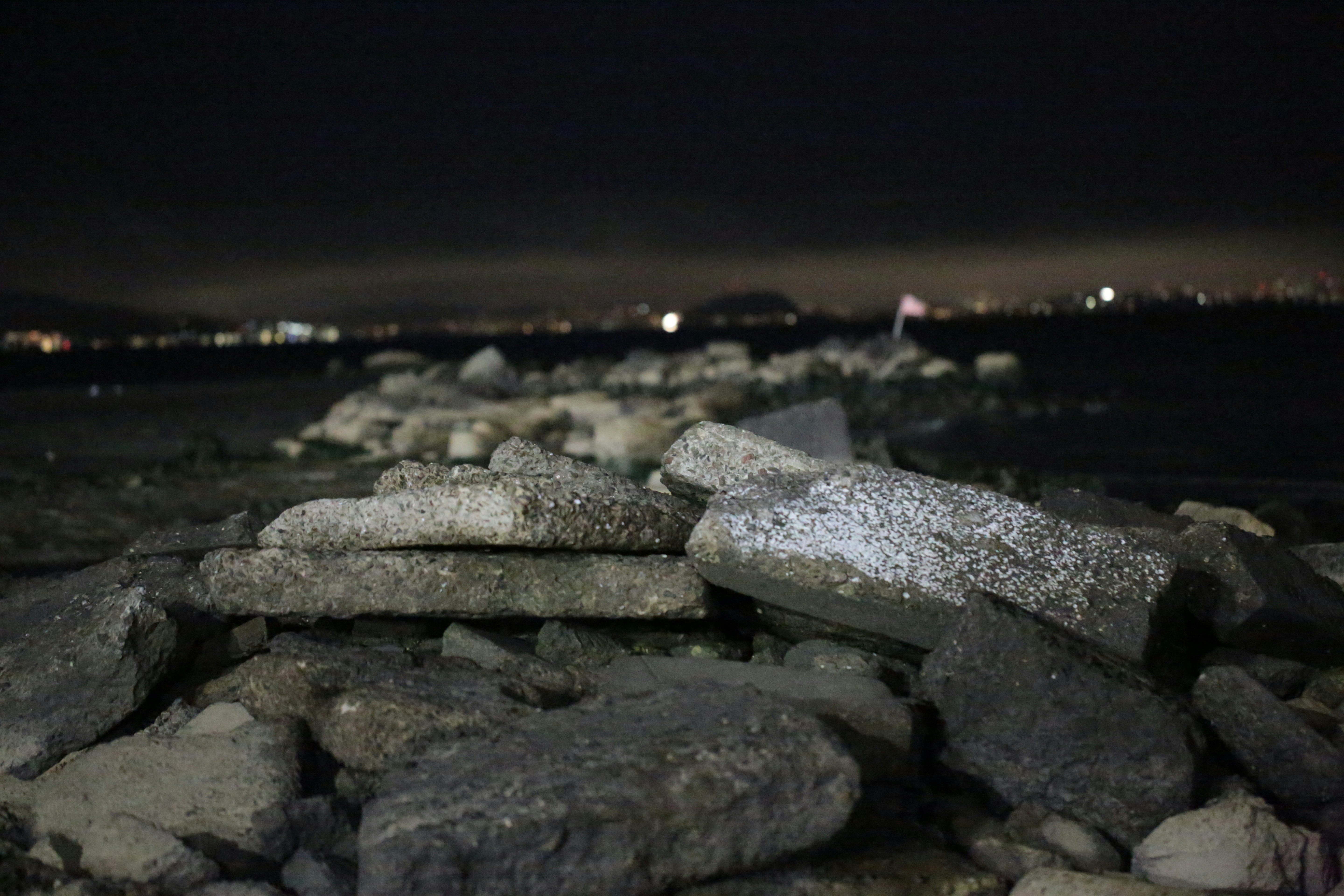 Stacked concrete slabs on a rocky shoreline at night with distant city lights