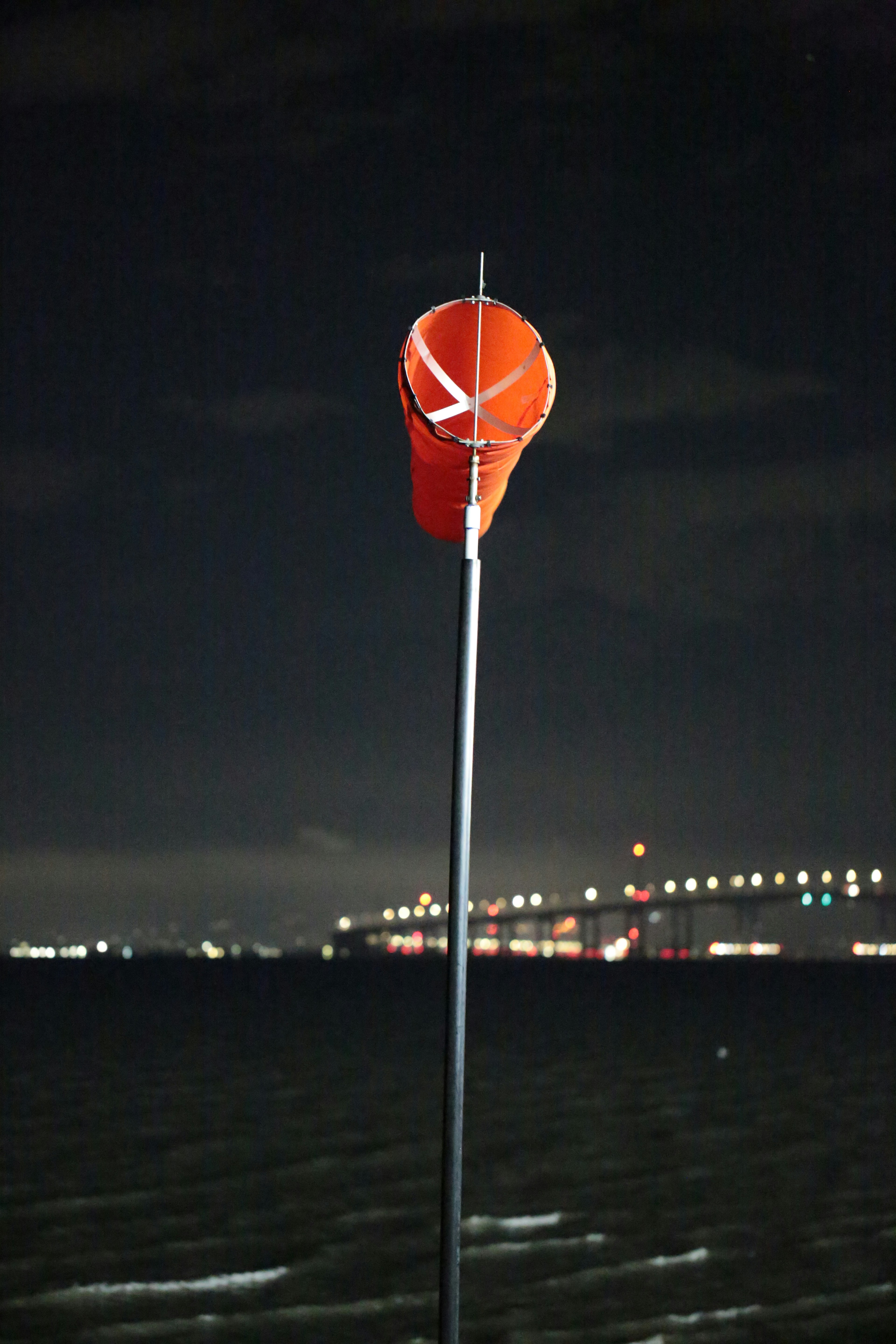 An orange windsock on a pole against a dark night sky with a distant bridge
