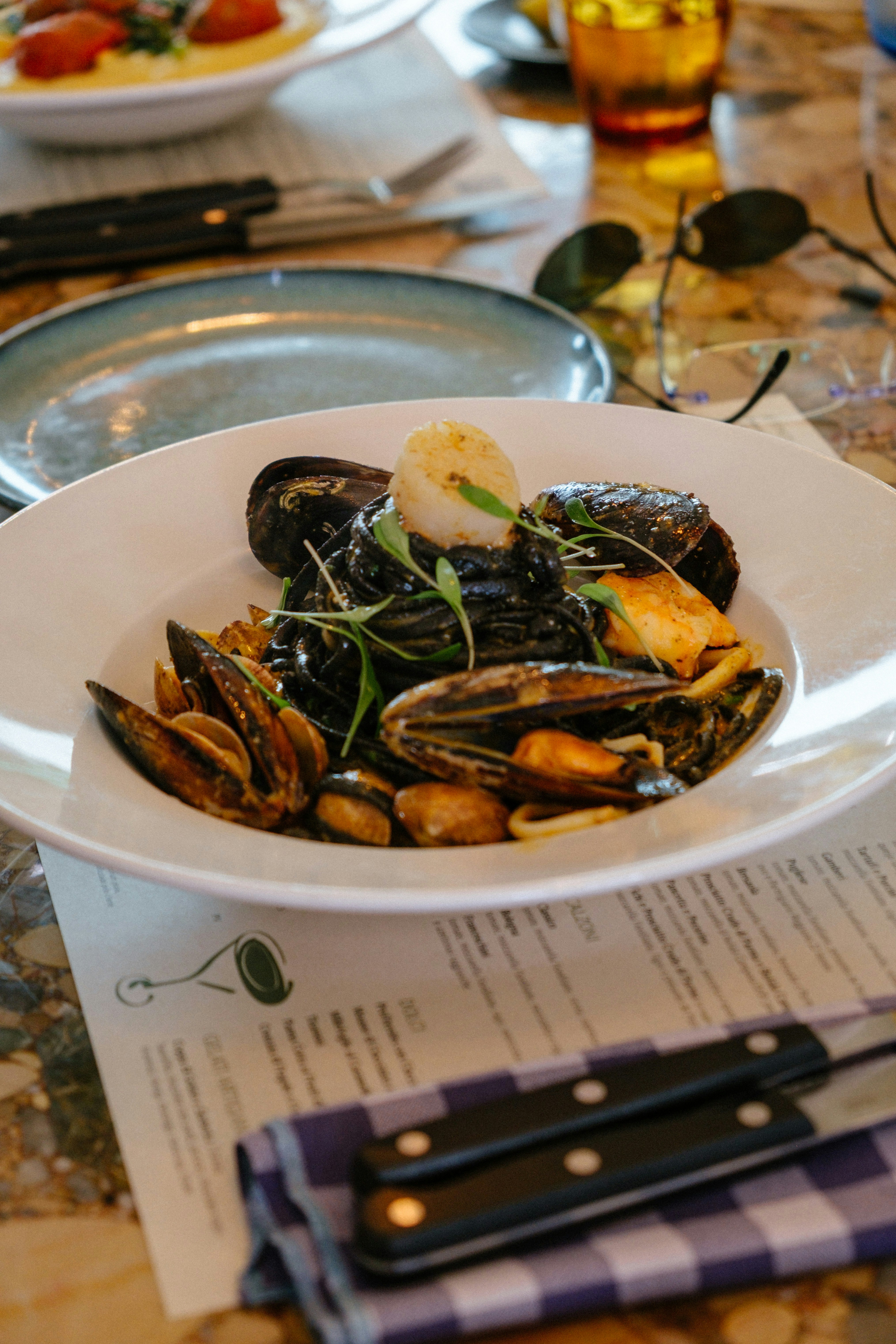 Black squid ink pasta with mussels, clams, and a scallop in a white bowl