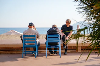 Three men sitting on blue chairs along a seaside promenade in warm light