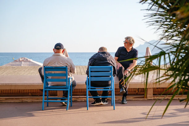 Three men sitting on blue chairs along a seaside promenade in warm light