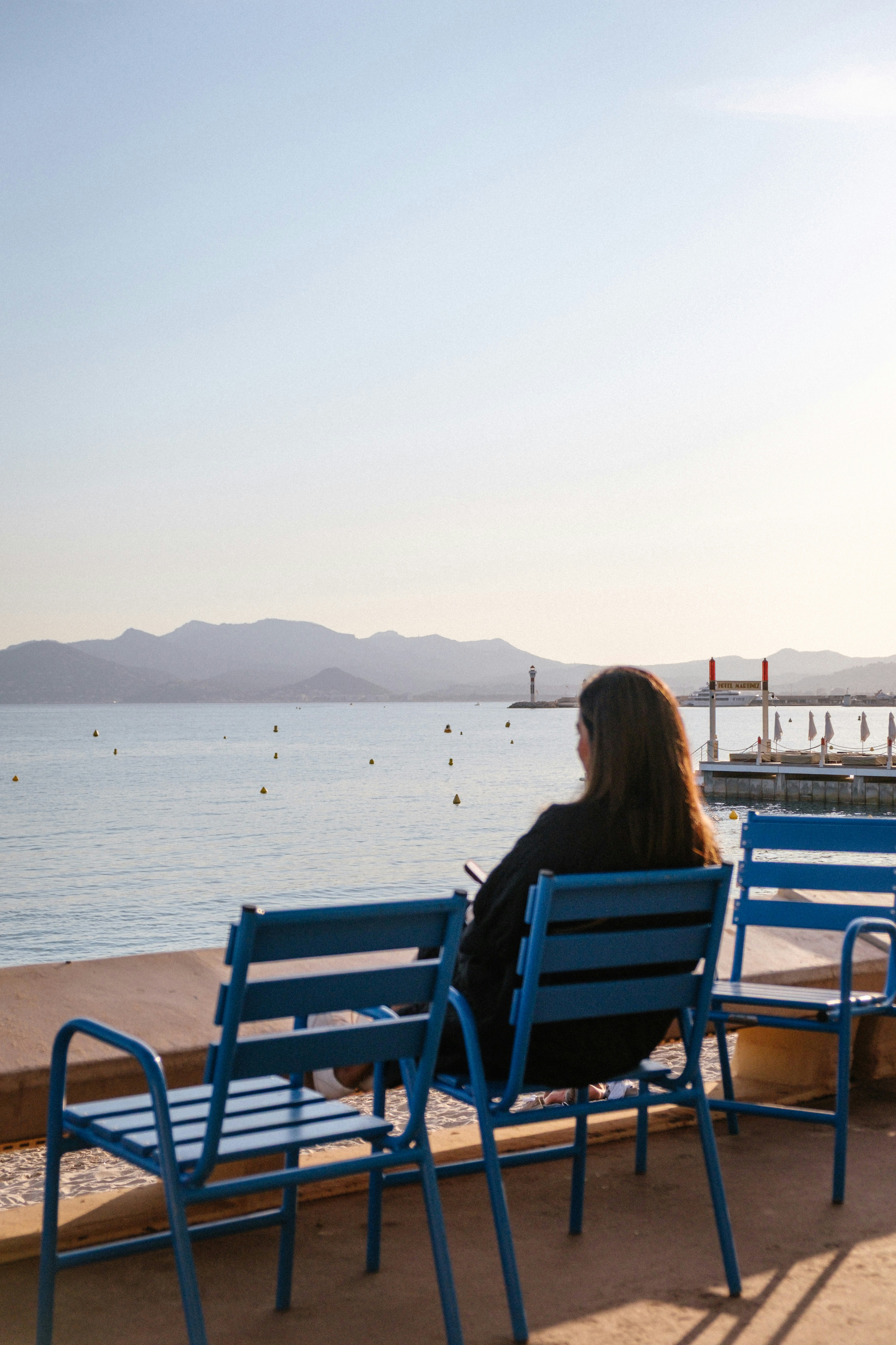 A person sitting on a blue chair facing the sea with distant mountains