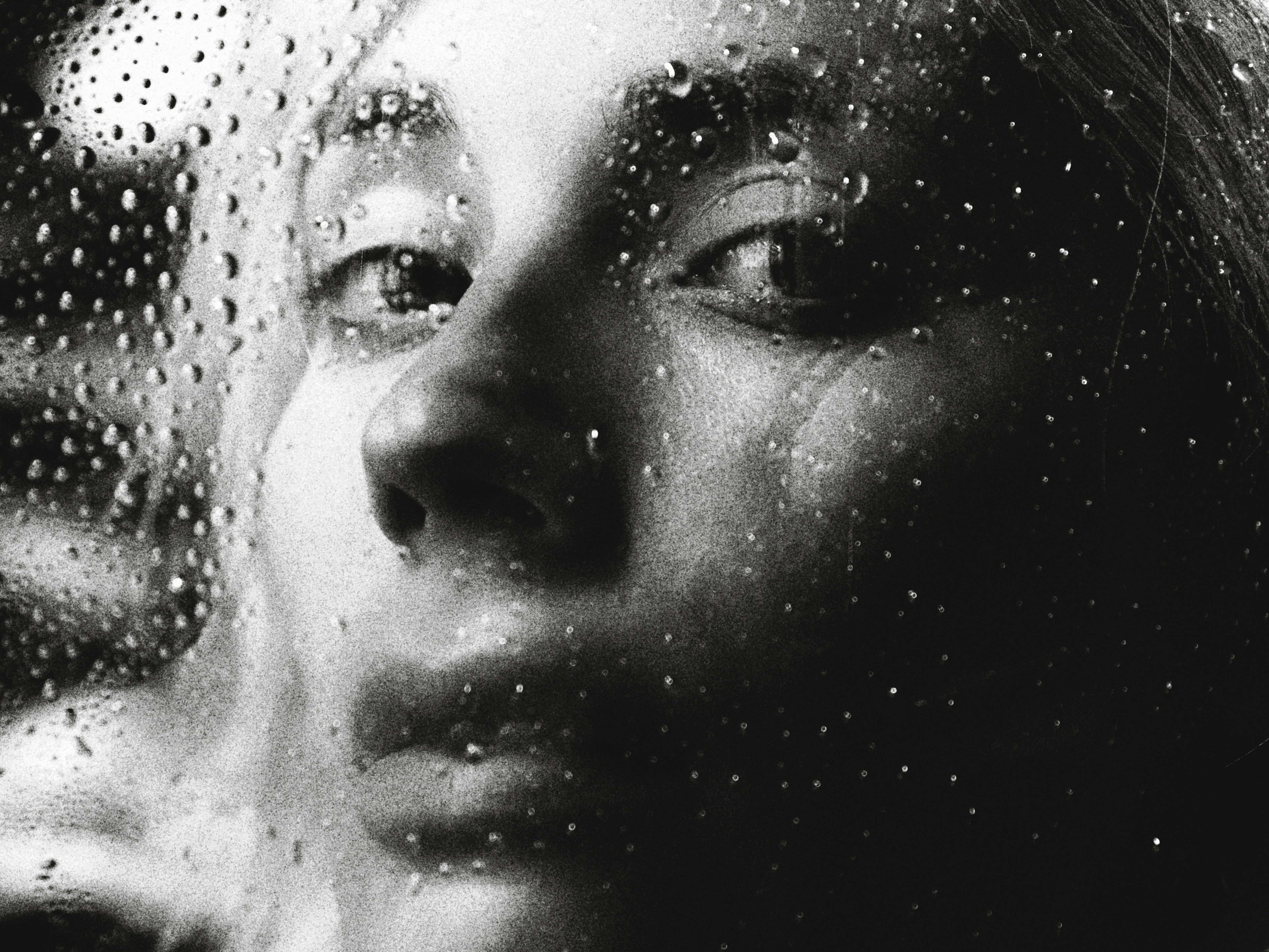 A black and white portrait of a person behind glass covered in water droplets