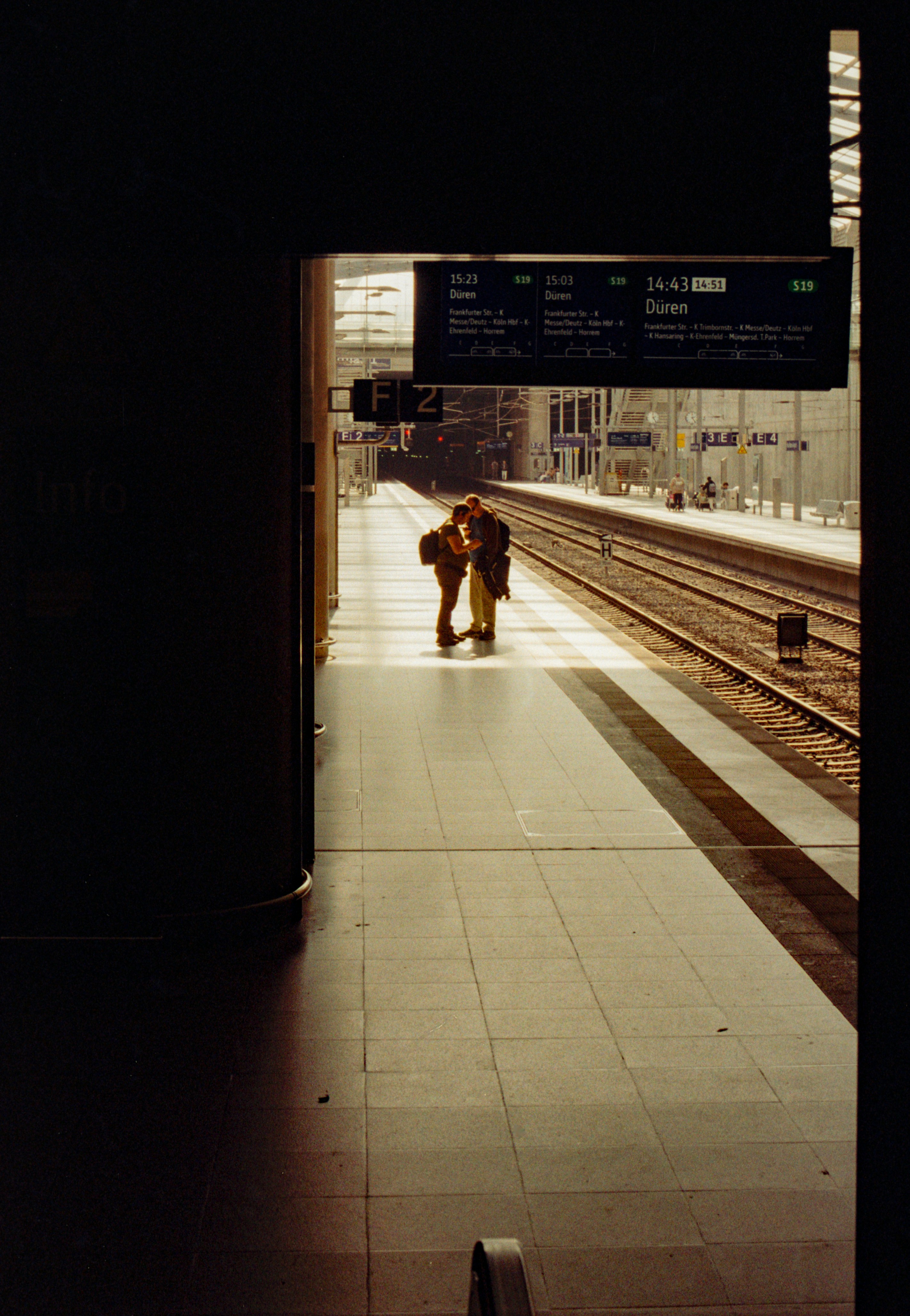 Zwei Personen mit Rucksäcken stehen auf einem Bahnsteig unter einer digitalen Abfahrtstafel