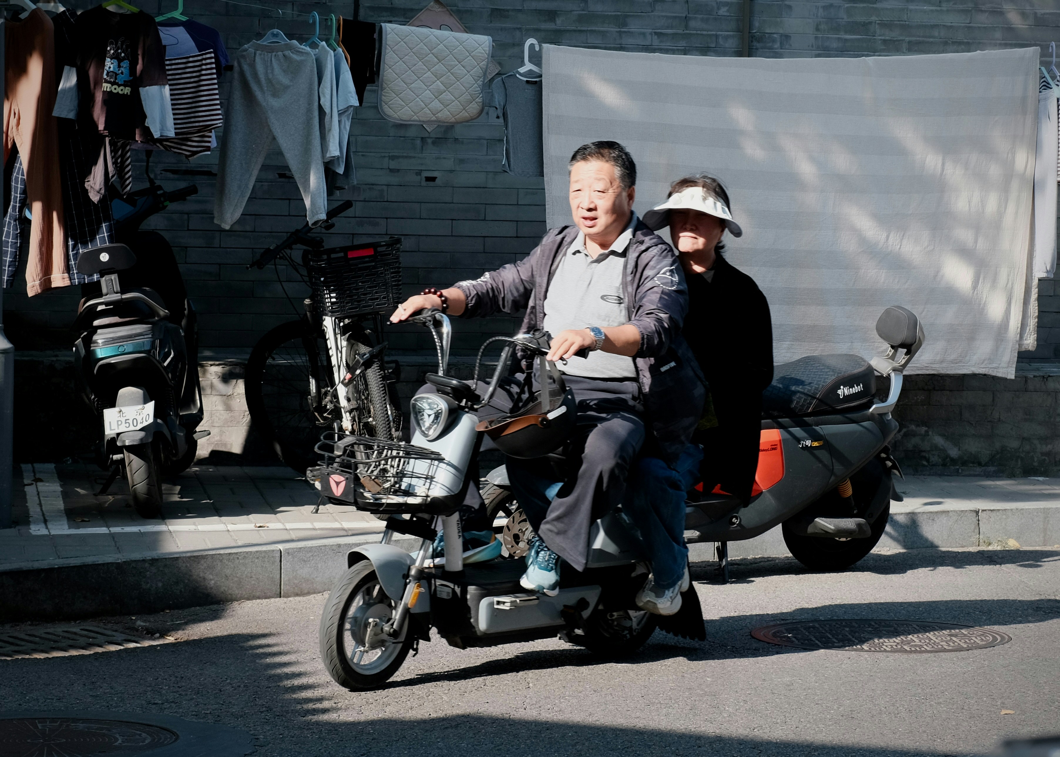 An adult man driving a scooter with a woman passenger on a sunny street