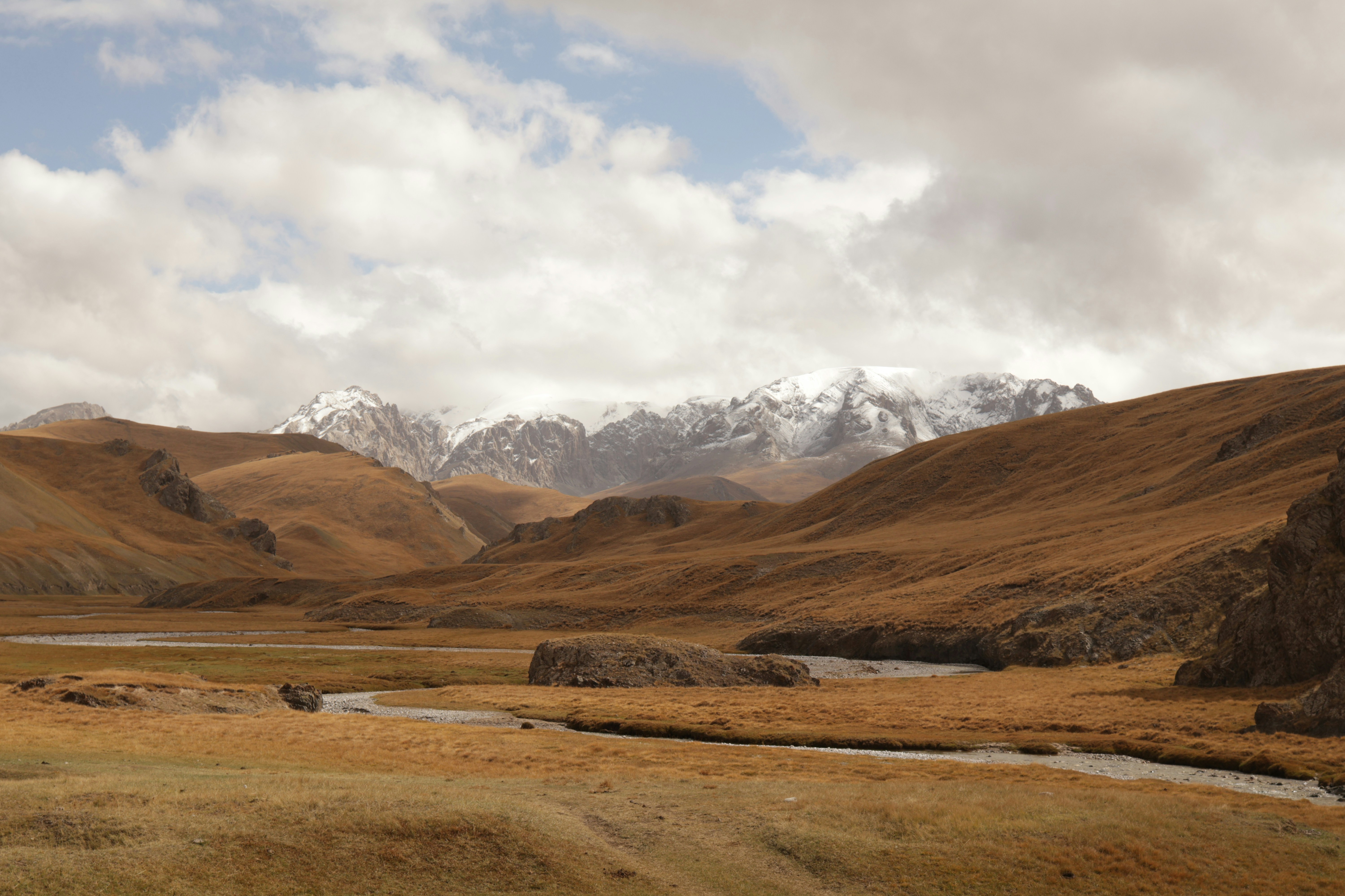 A winding river through a golden valley beneath snow-capped mountains under a cloudy sky