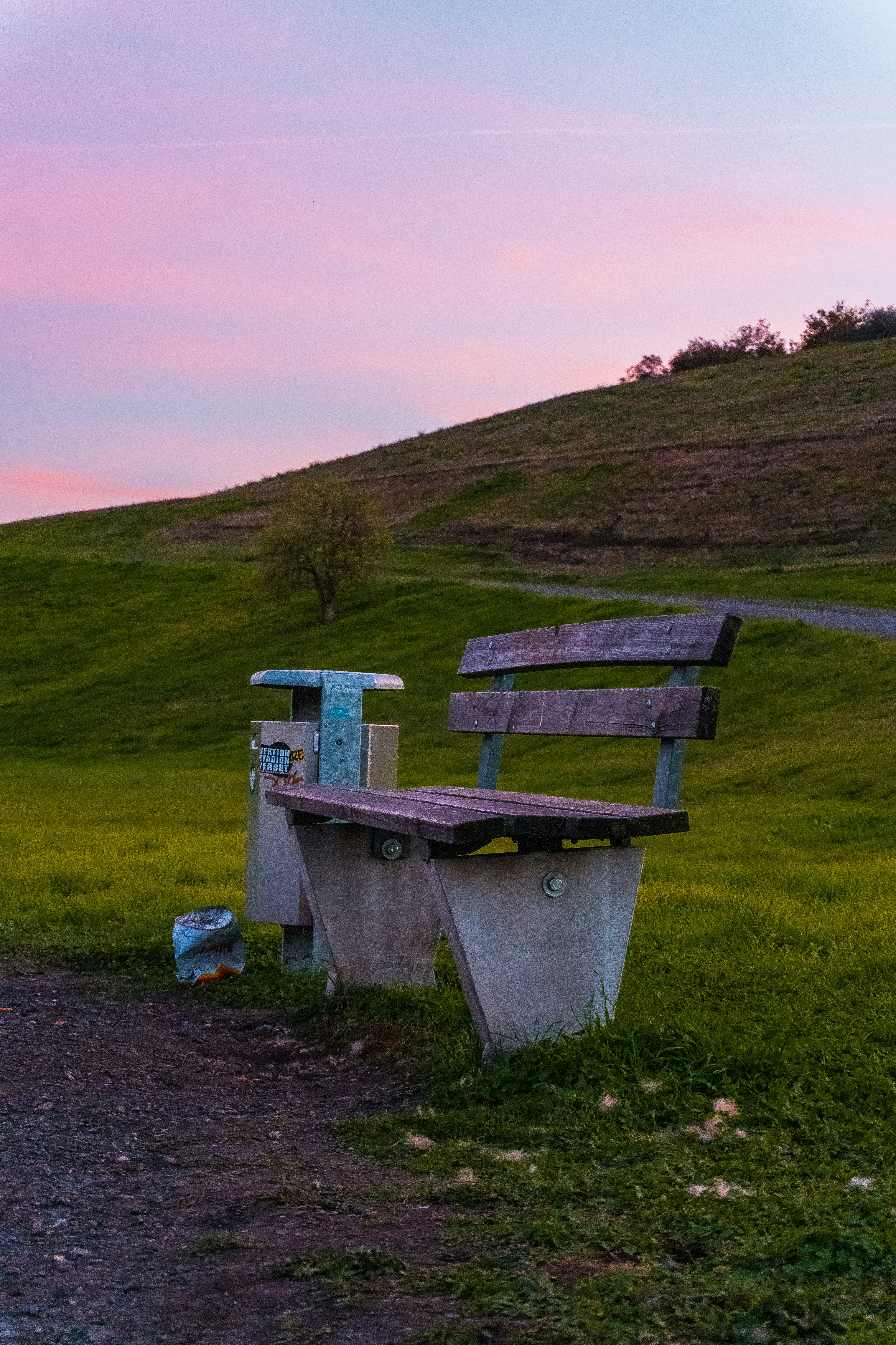 A wooden bench and a trash bin on a grassy hill under a pink sky