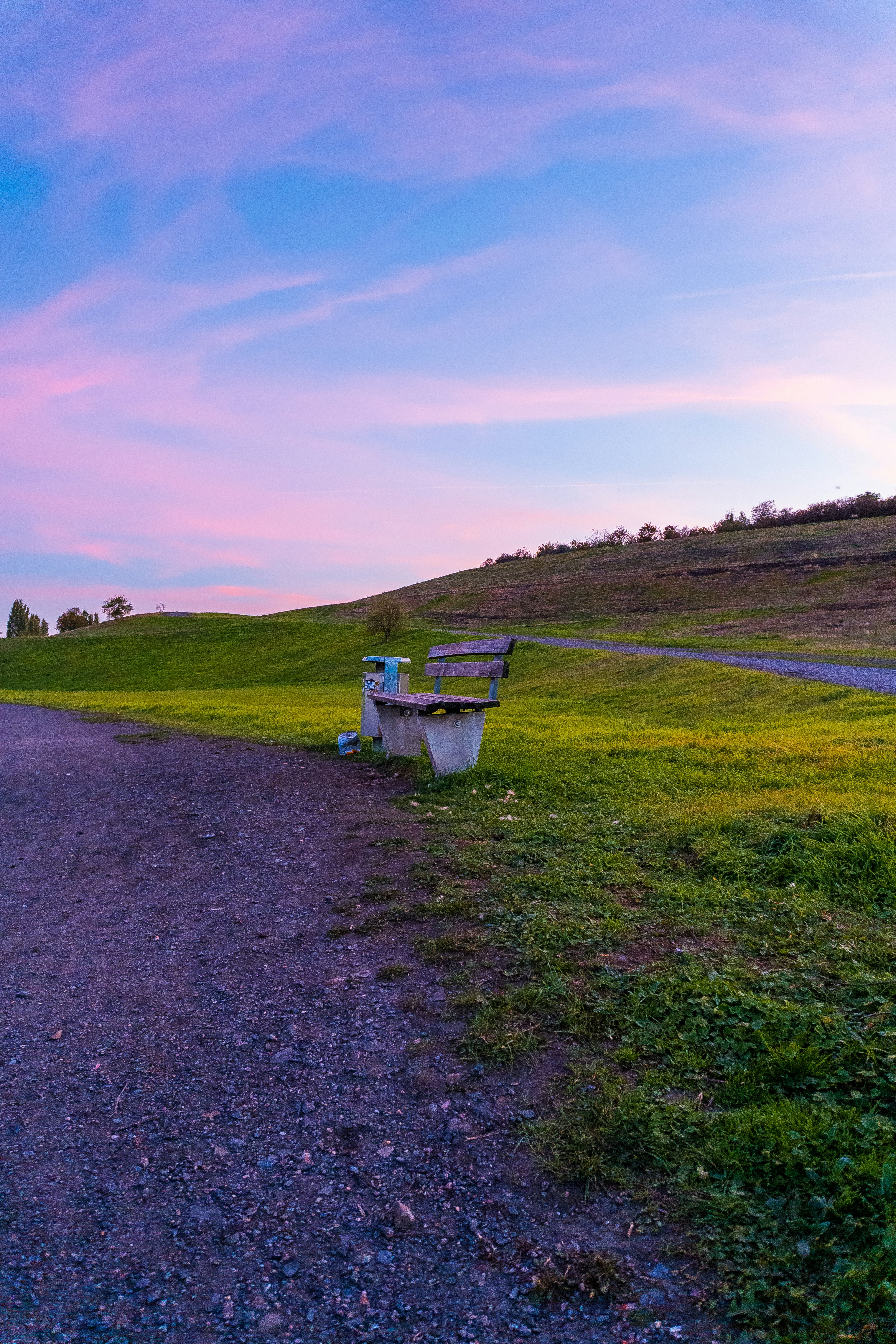 A wooden bench beside a trash bin on a grassy hill under a pink and blue sky