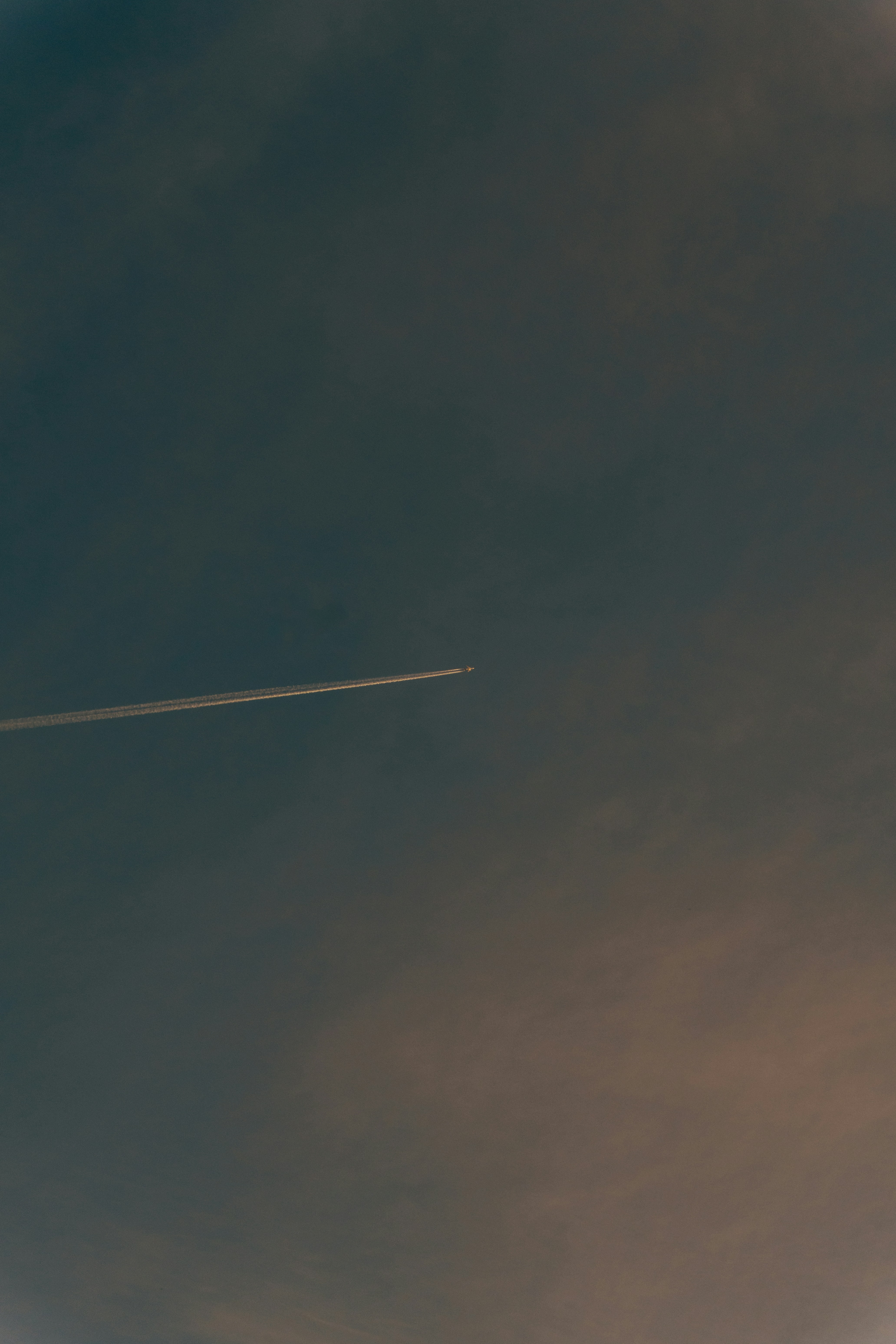 A distant airplane leaving a long white contrail across a dark moody sky