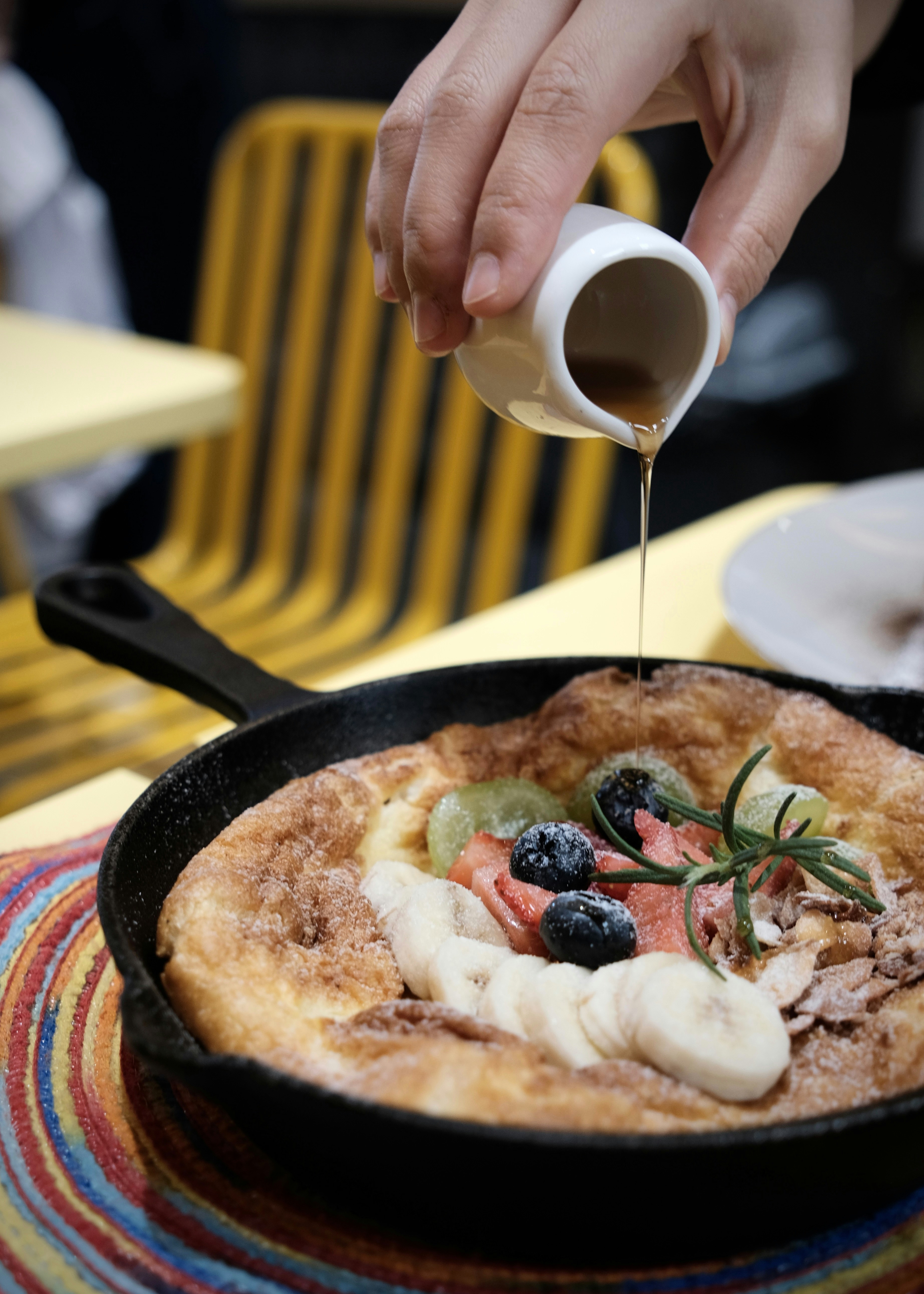 Syrup pouring from a small white pitcher onto a fruit-topped Dutch baby pancake in a cast iron skillet