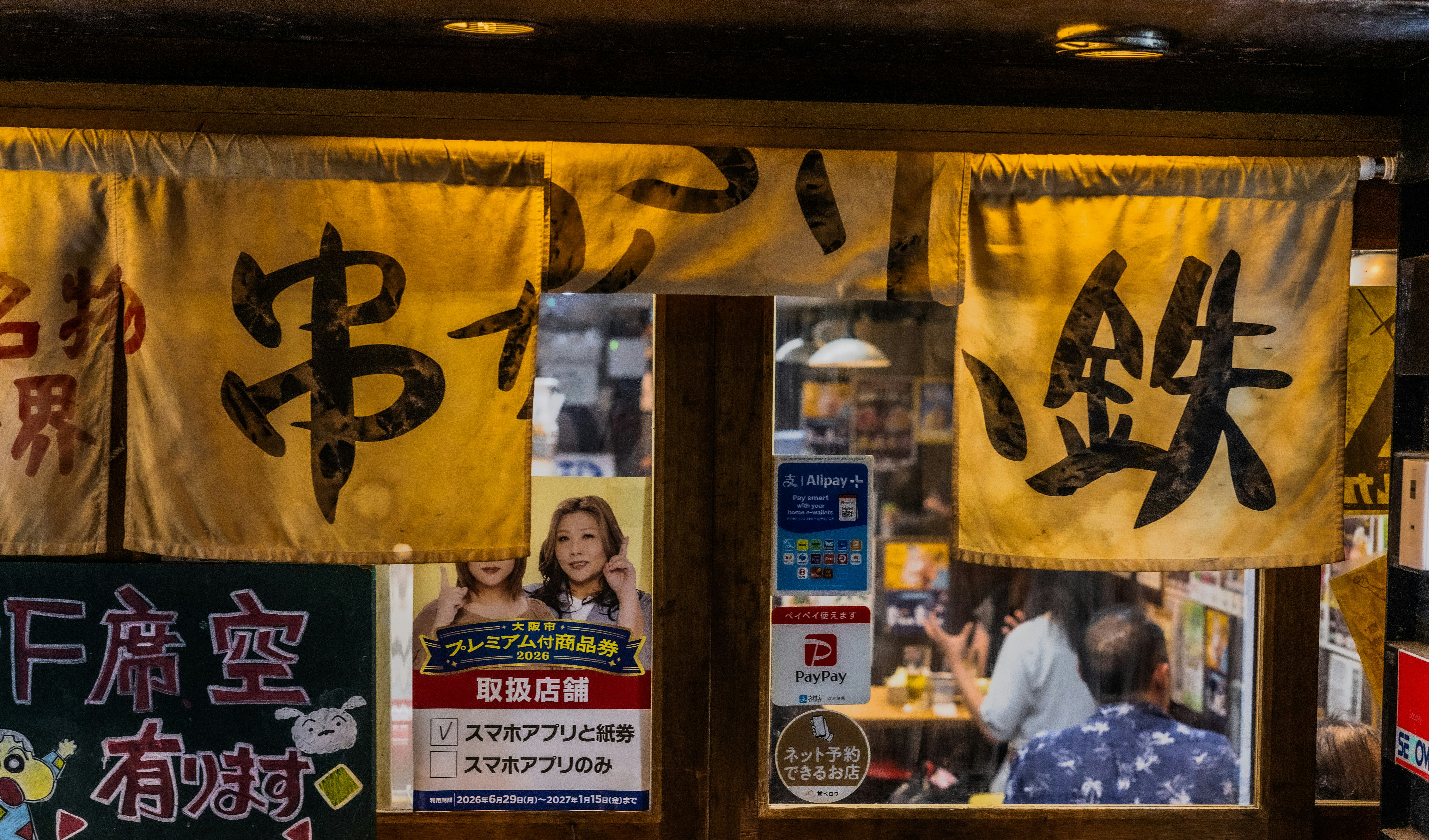 A restaurant entrance with yellow fabric curtains featuring black Japanese calligraphy and various payment method stickers