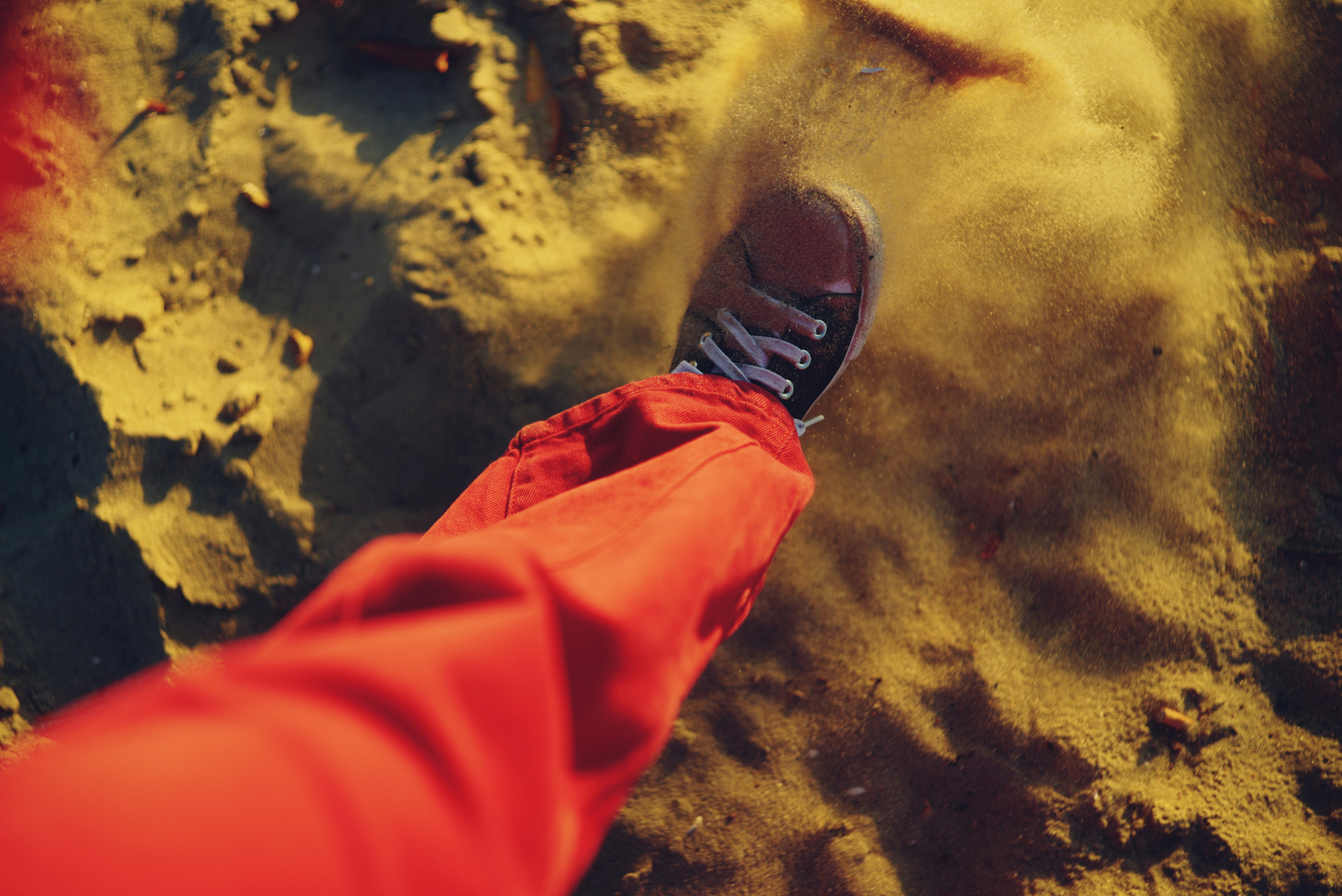 Une personne en pantalon rouge soulevant du sable avec une basket dans une lumière dorée et chaude