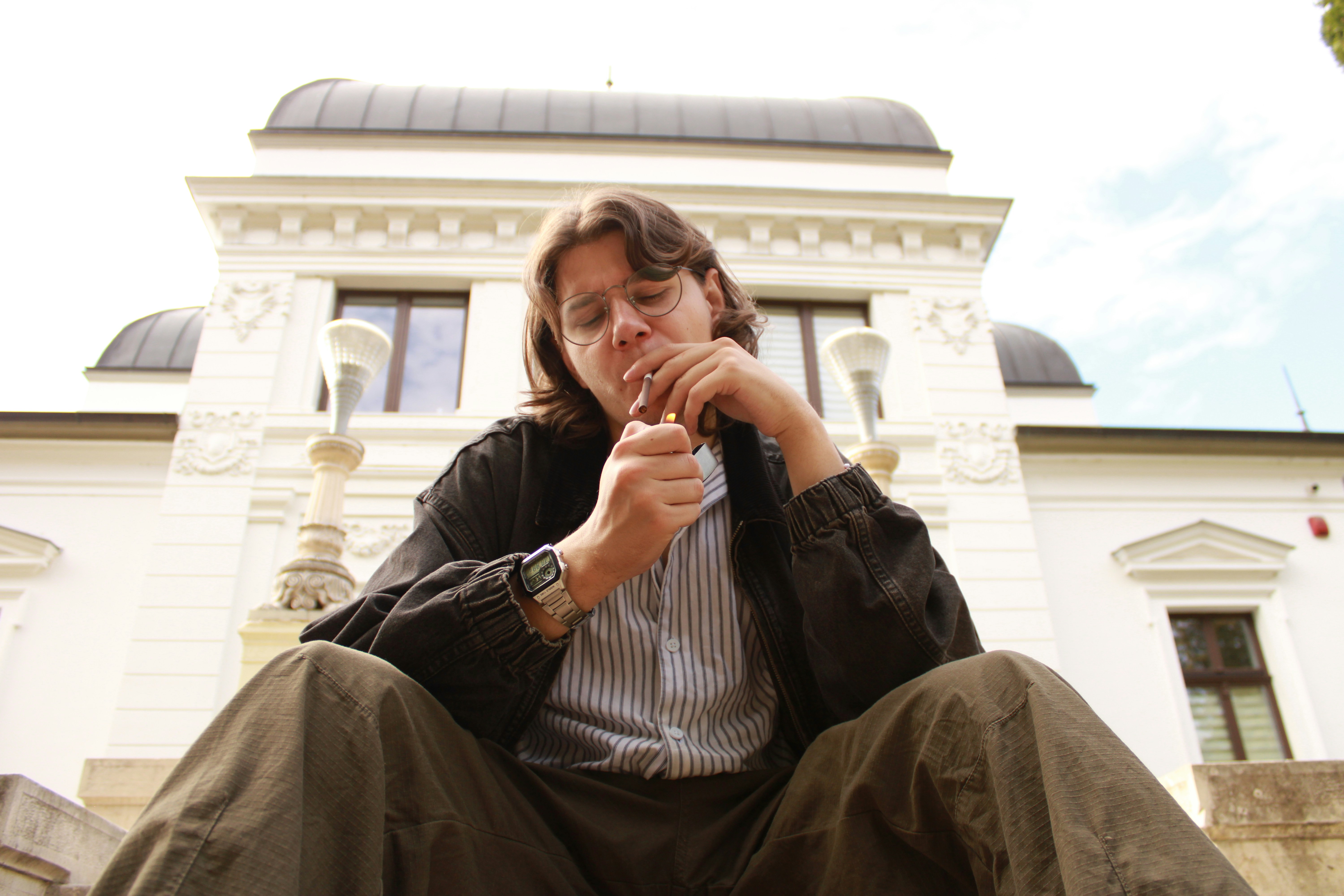 A person with glasses lighting a cigarette in front of a white neoclassical building