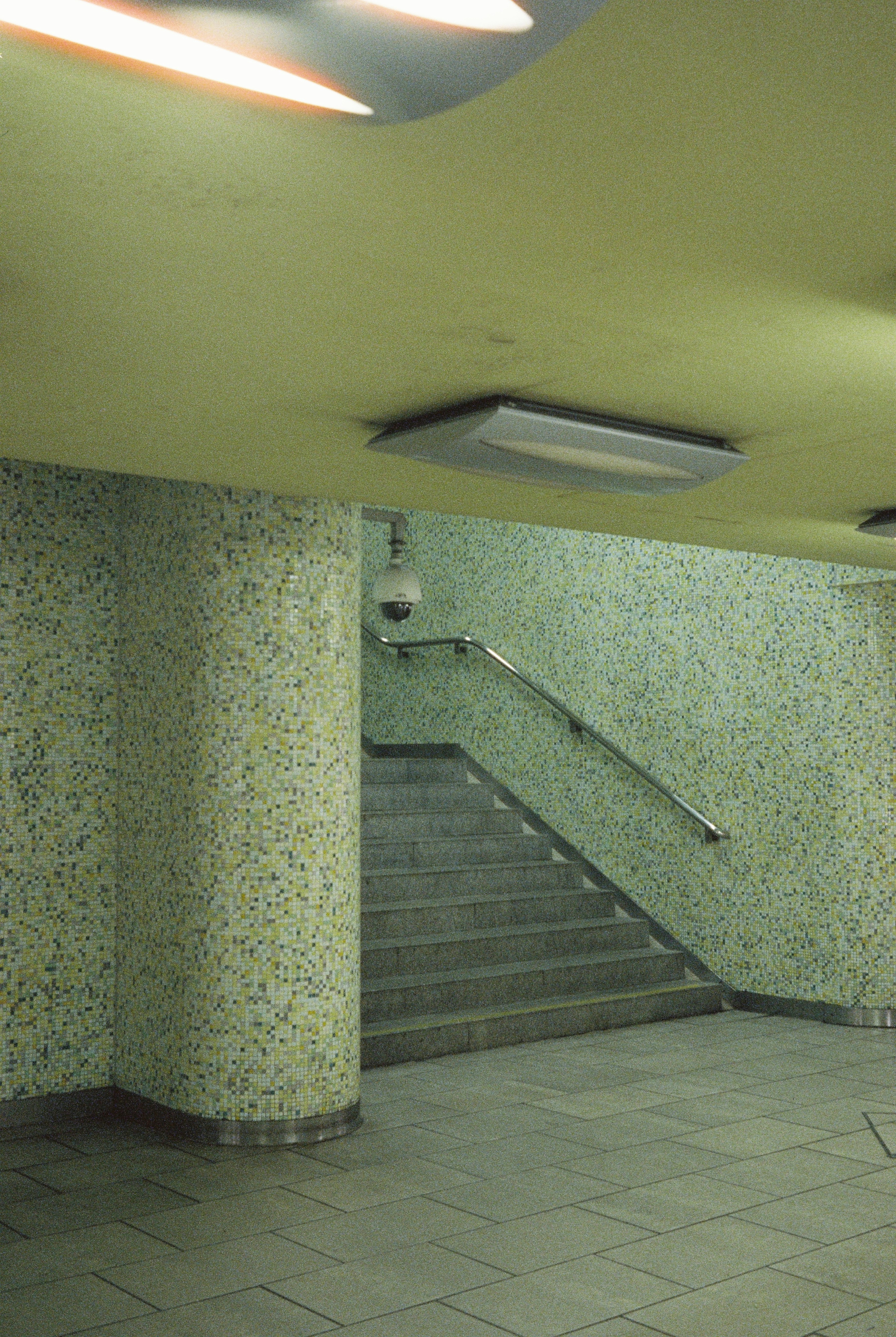A staircase with a metal handrail leading up between walls covered in small yellow and green mosaic tiles