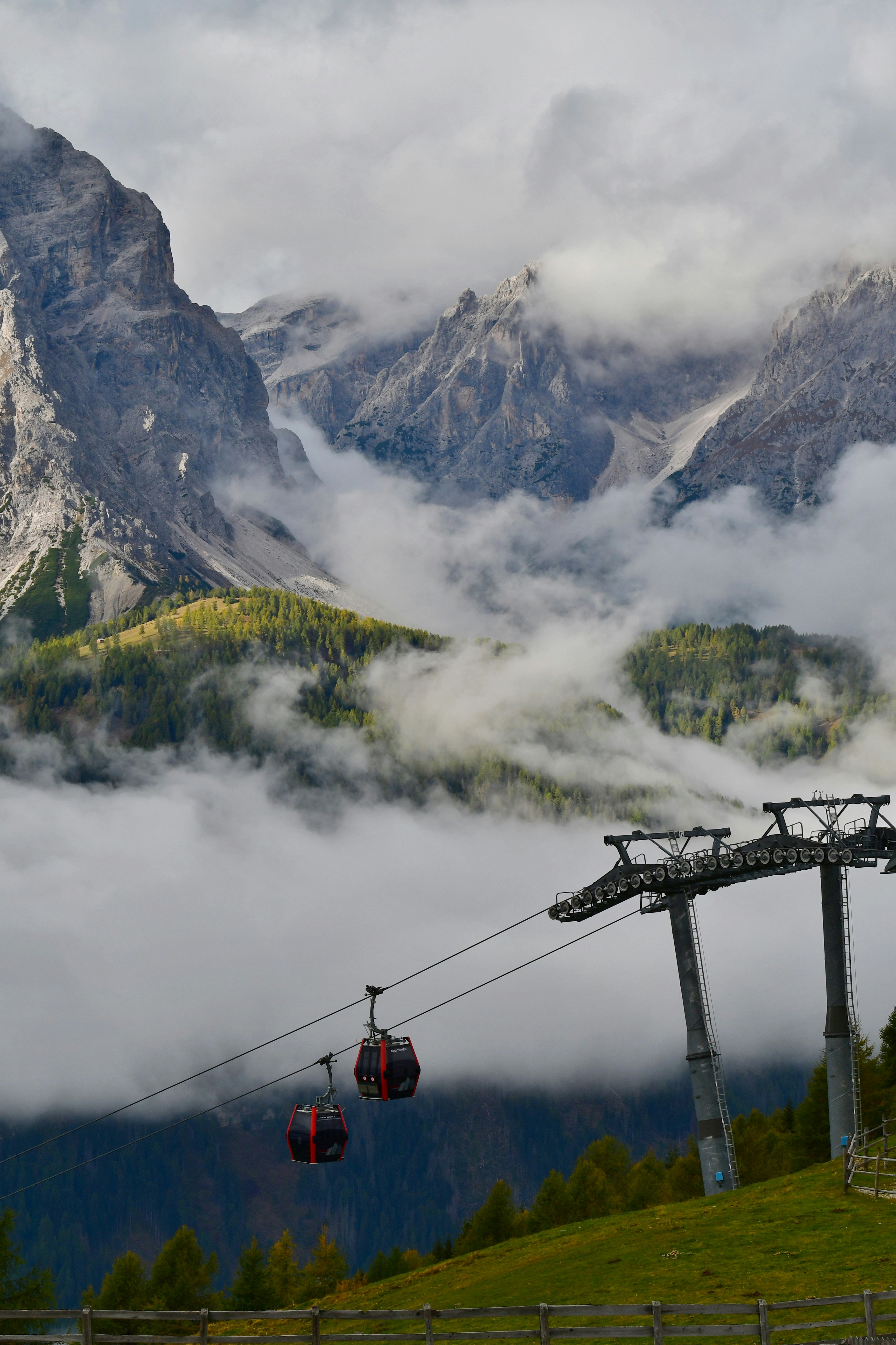 Tranvías de cable rojos avanzando entre densas nubes sobre una ladera verde de montaña con picos rocosos