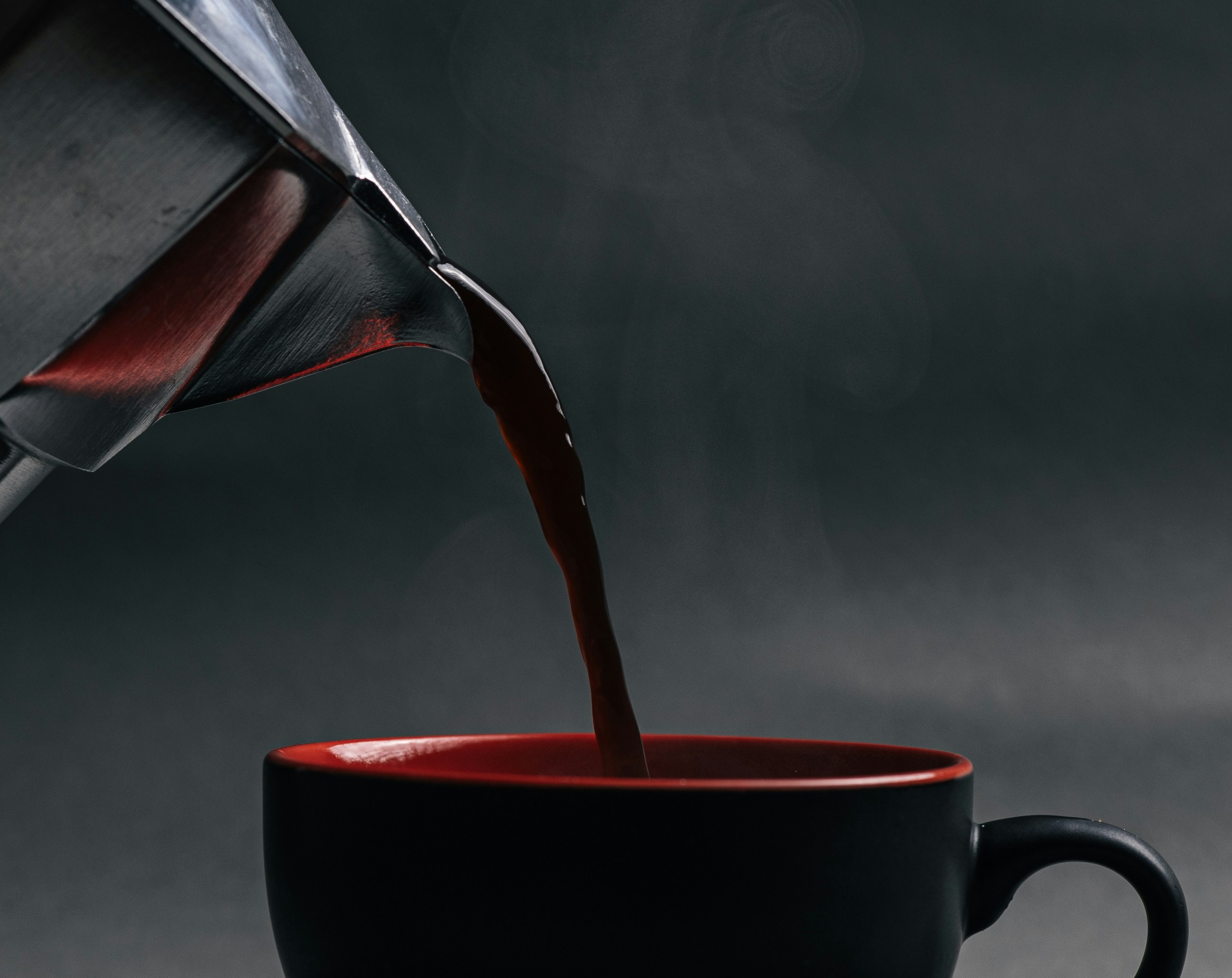 Coffee pouring from a metal moka pot into a black and red ceramic cup