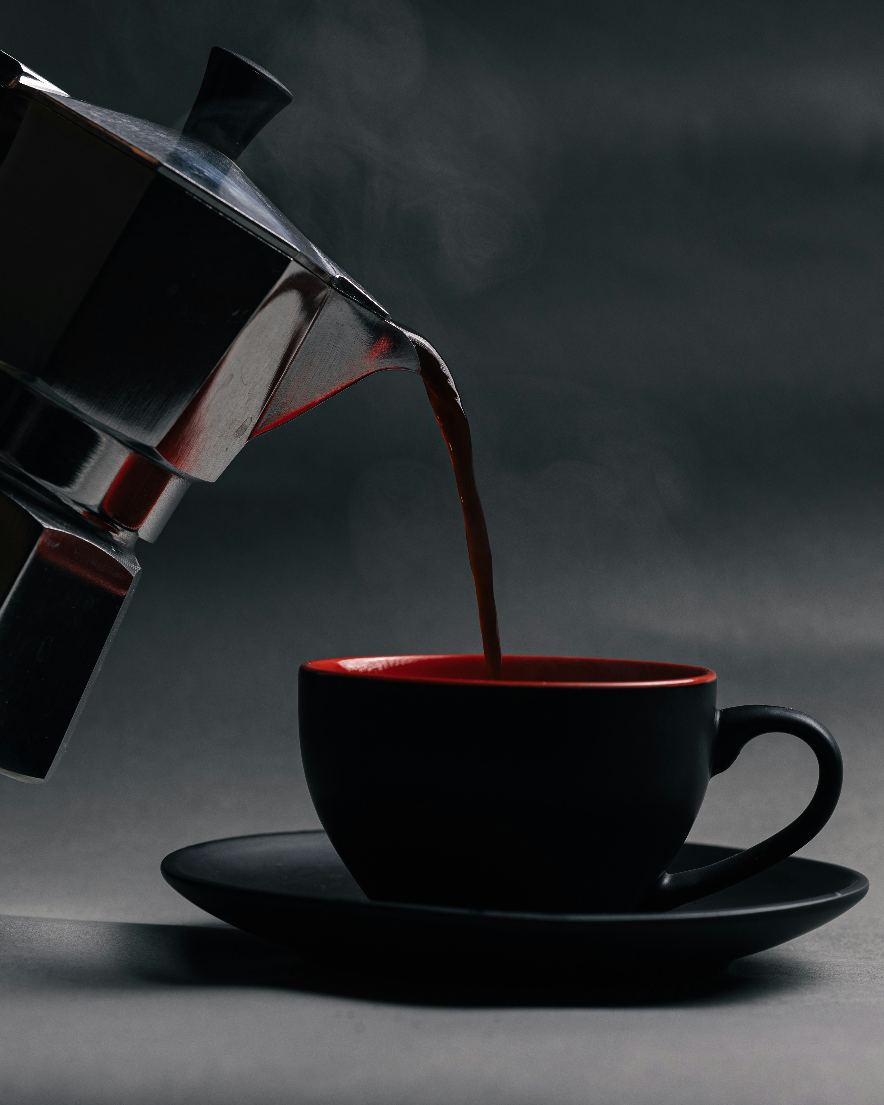 Coffee pouring from a metallic moka pot into a black and red cup