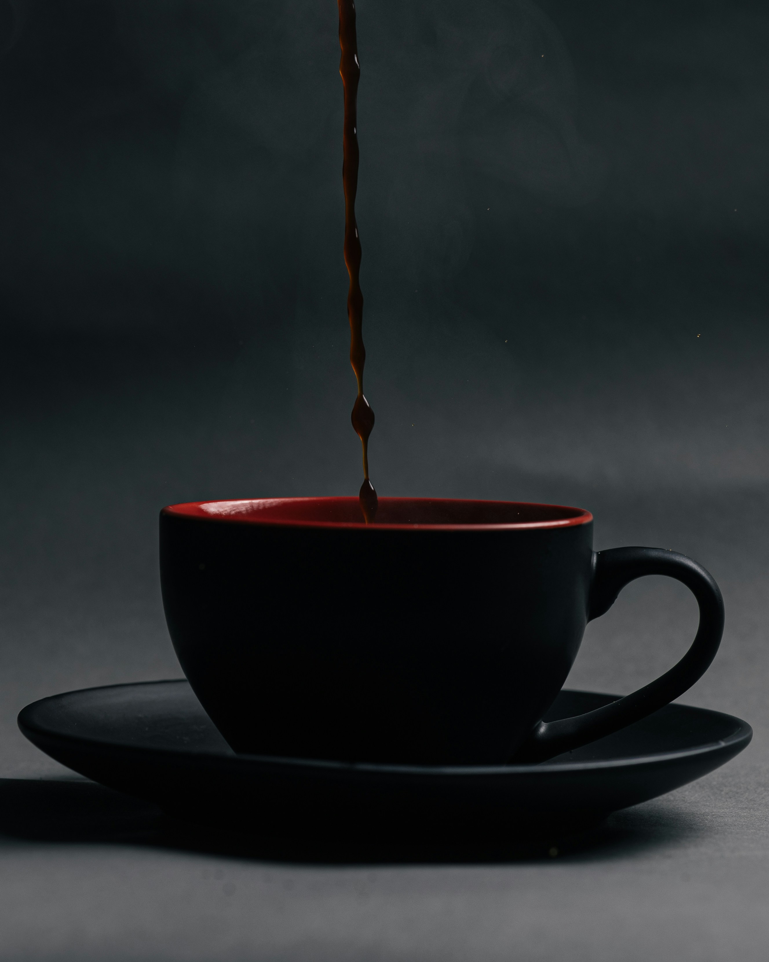Dark coffee pouring into a black and red ceramic cup on a saucer