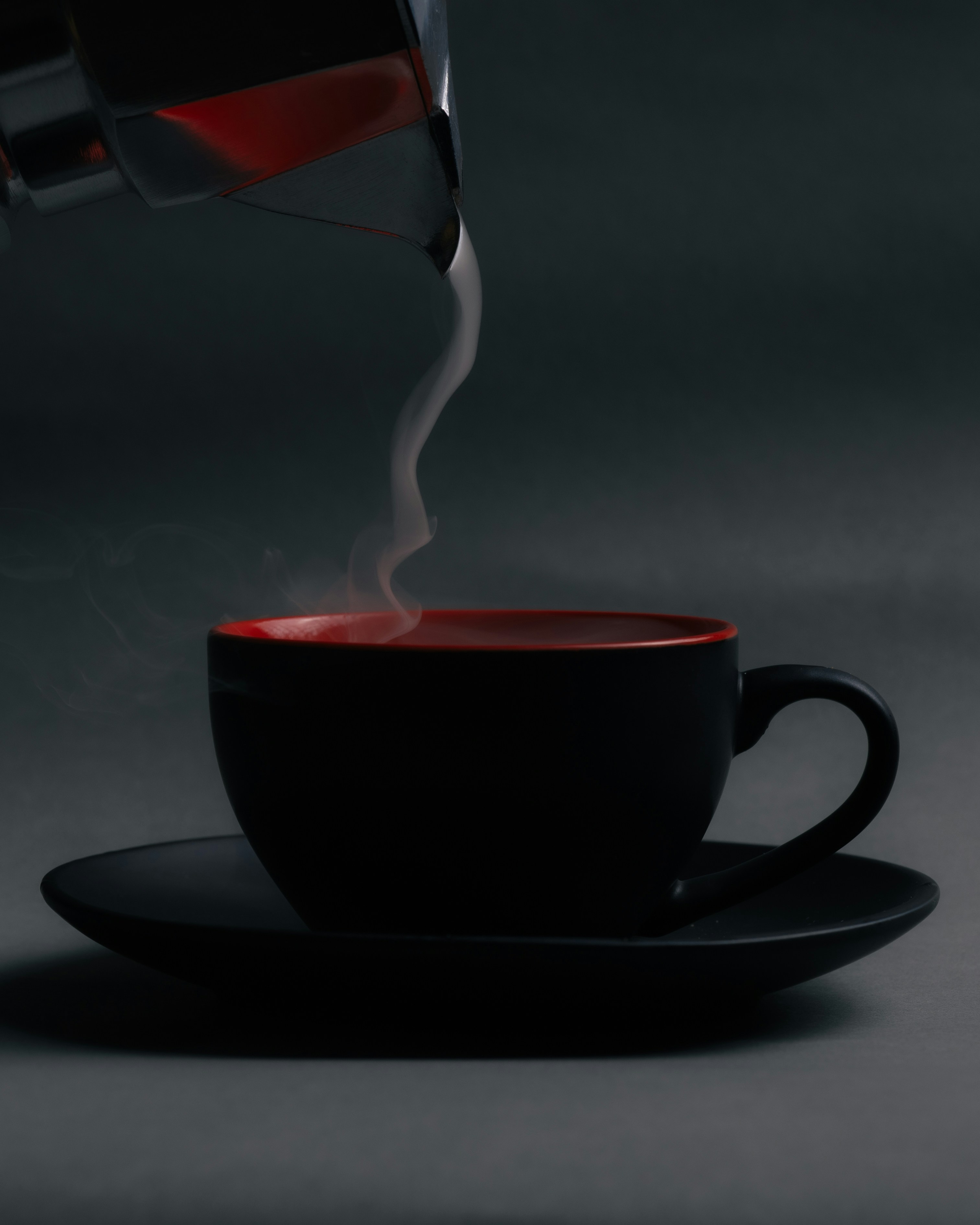 Hot liquid pouring from a metal pot into a black and red coffee cup