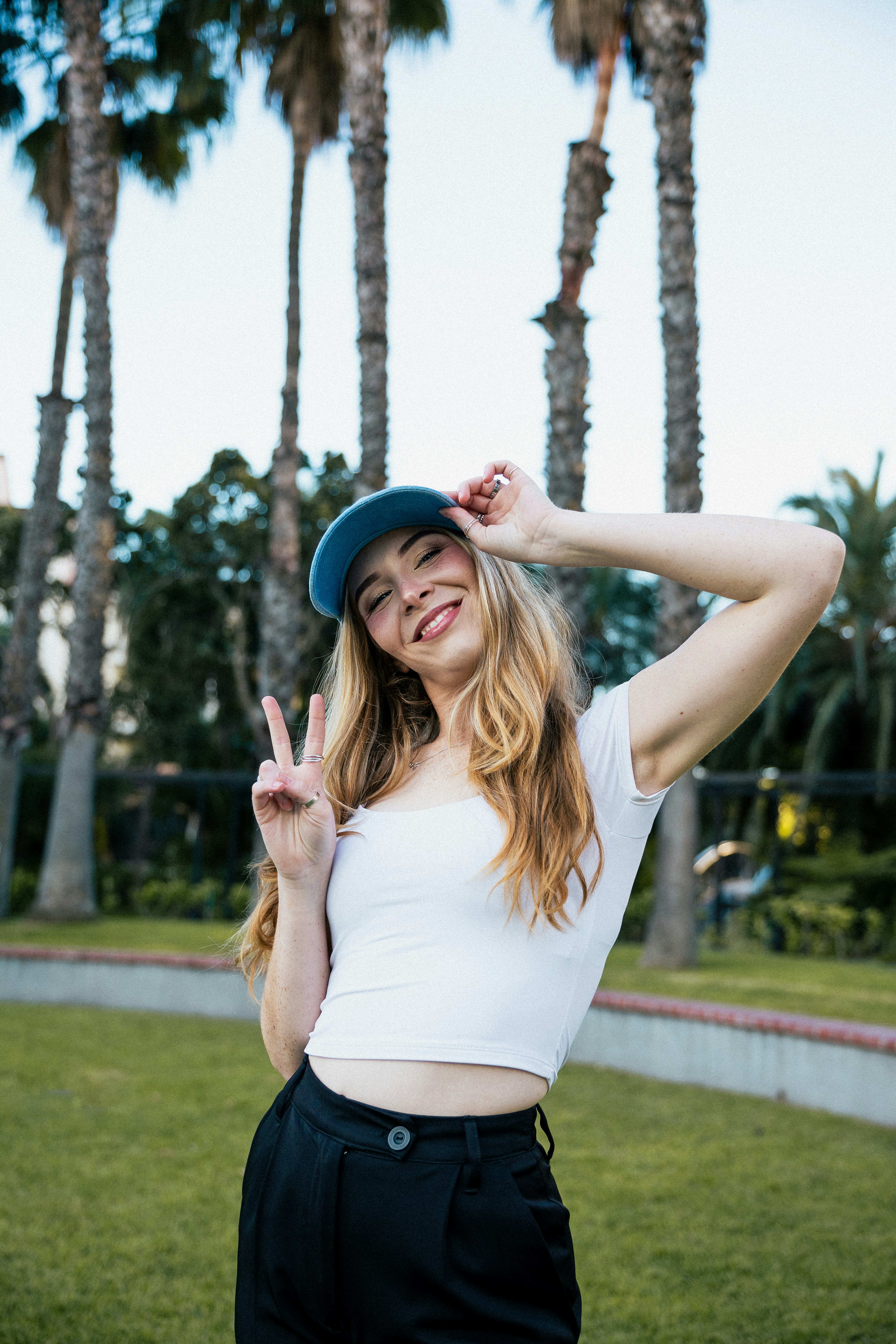 Seorang wanita muda dengan crop top putih dan topi biru membuat tanda perdamaian di taman