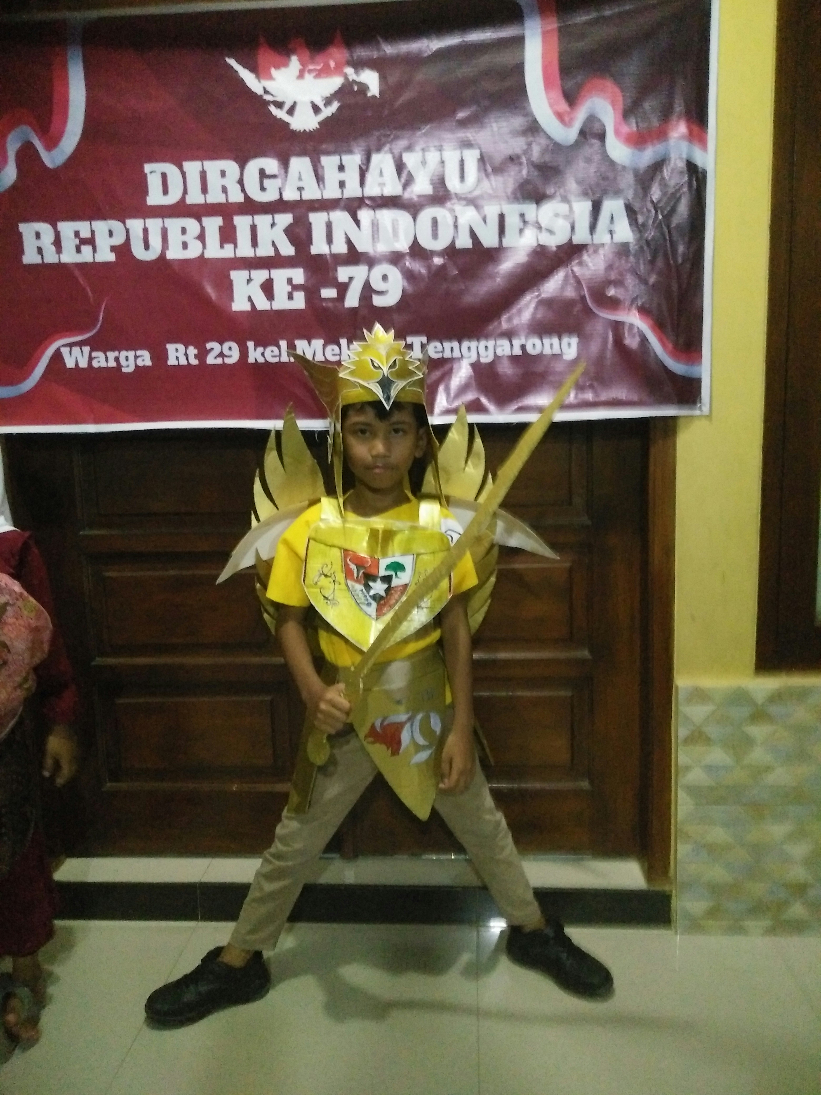 Seorang anak kecil dengan kostum Garuda emas memegang pedang di depan spanduk kemerdekaan Indonesia
