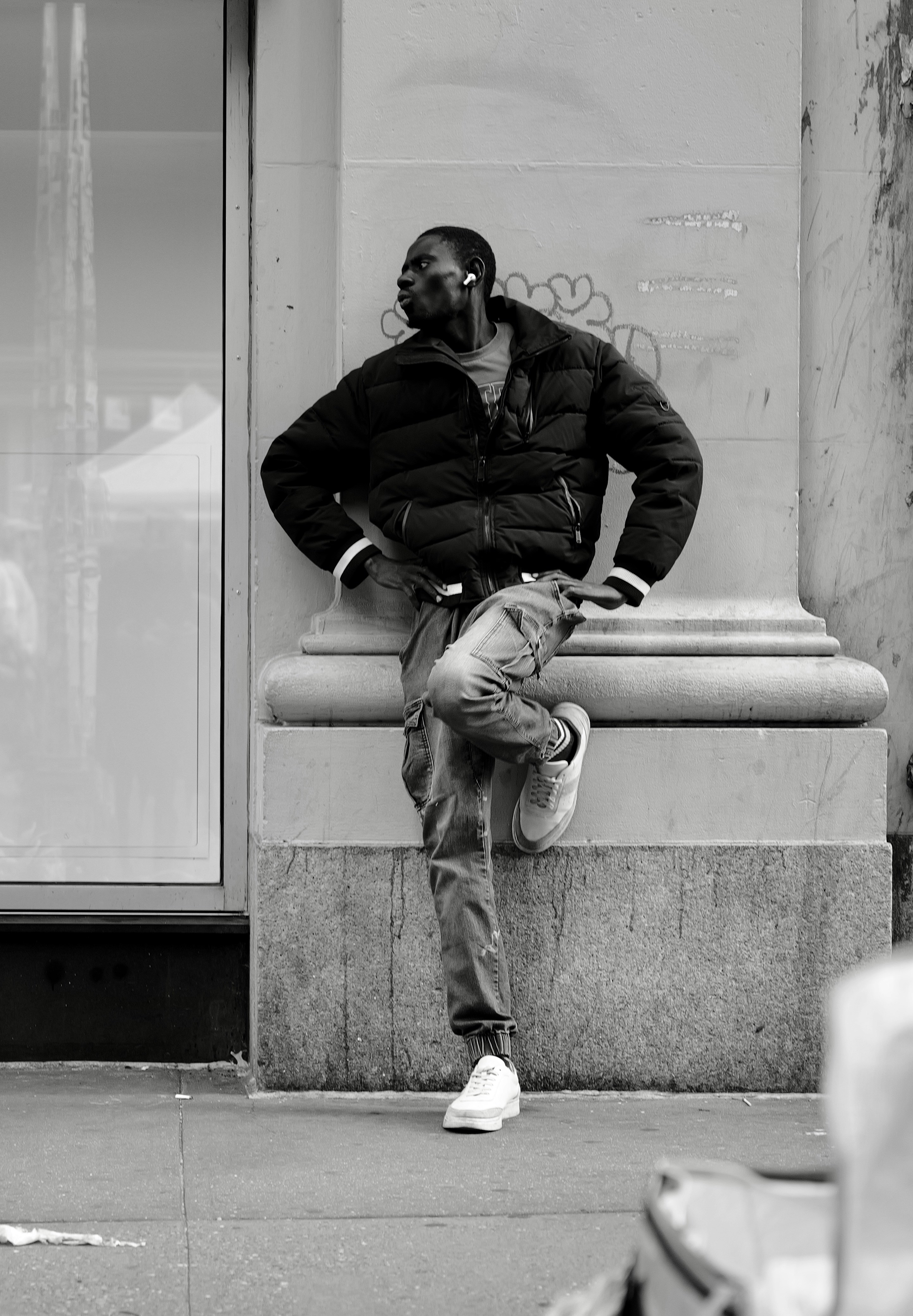 A man in a black puffer jacket and jeans leaning against a stone wall