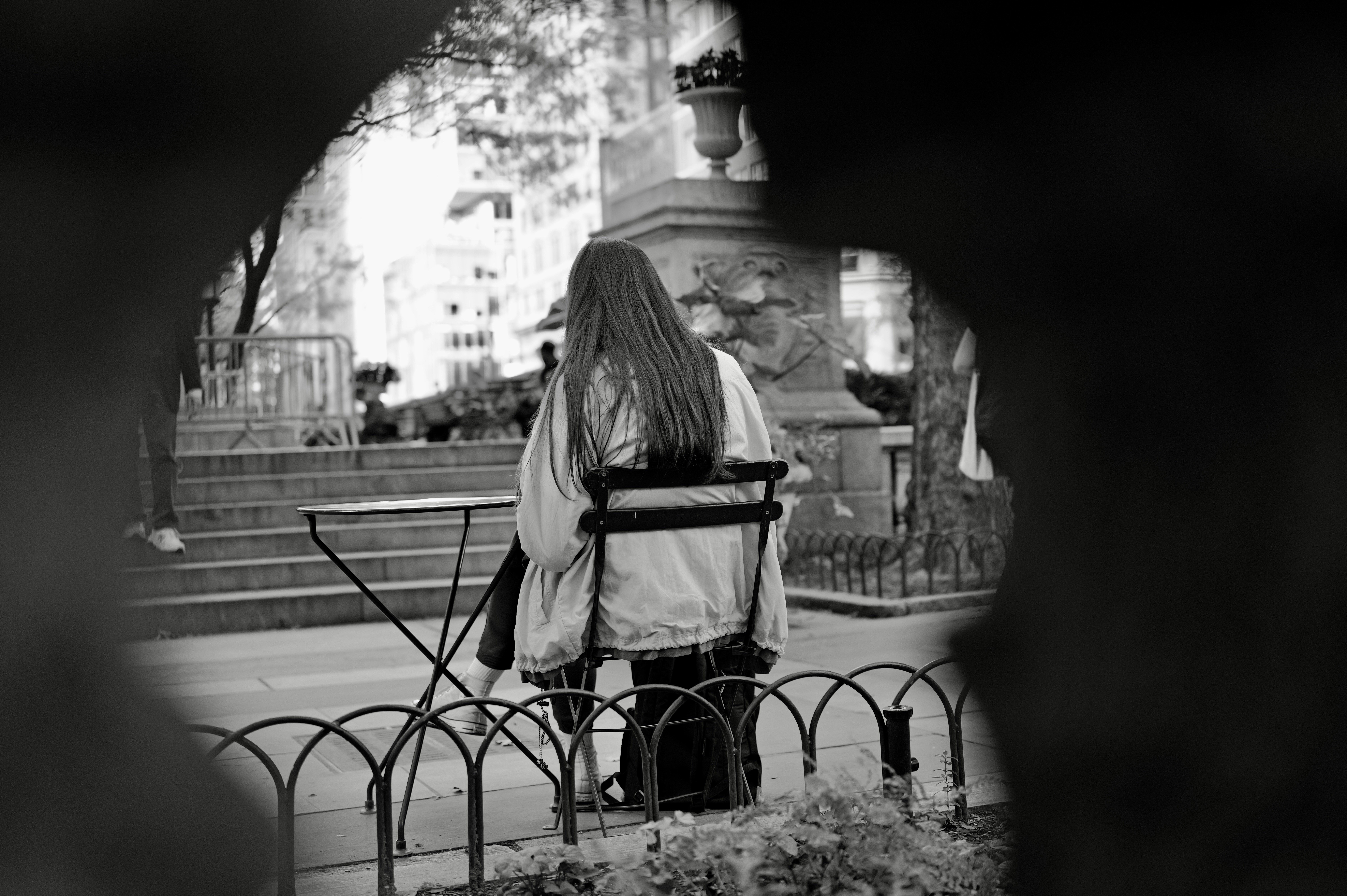 A person with long hair sitting at a small table in a park, black and white