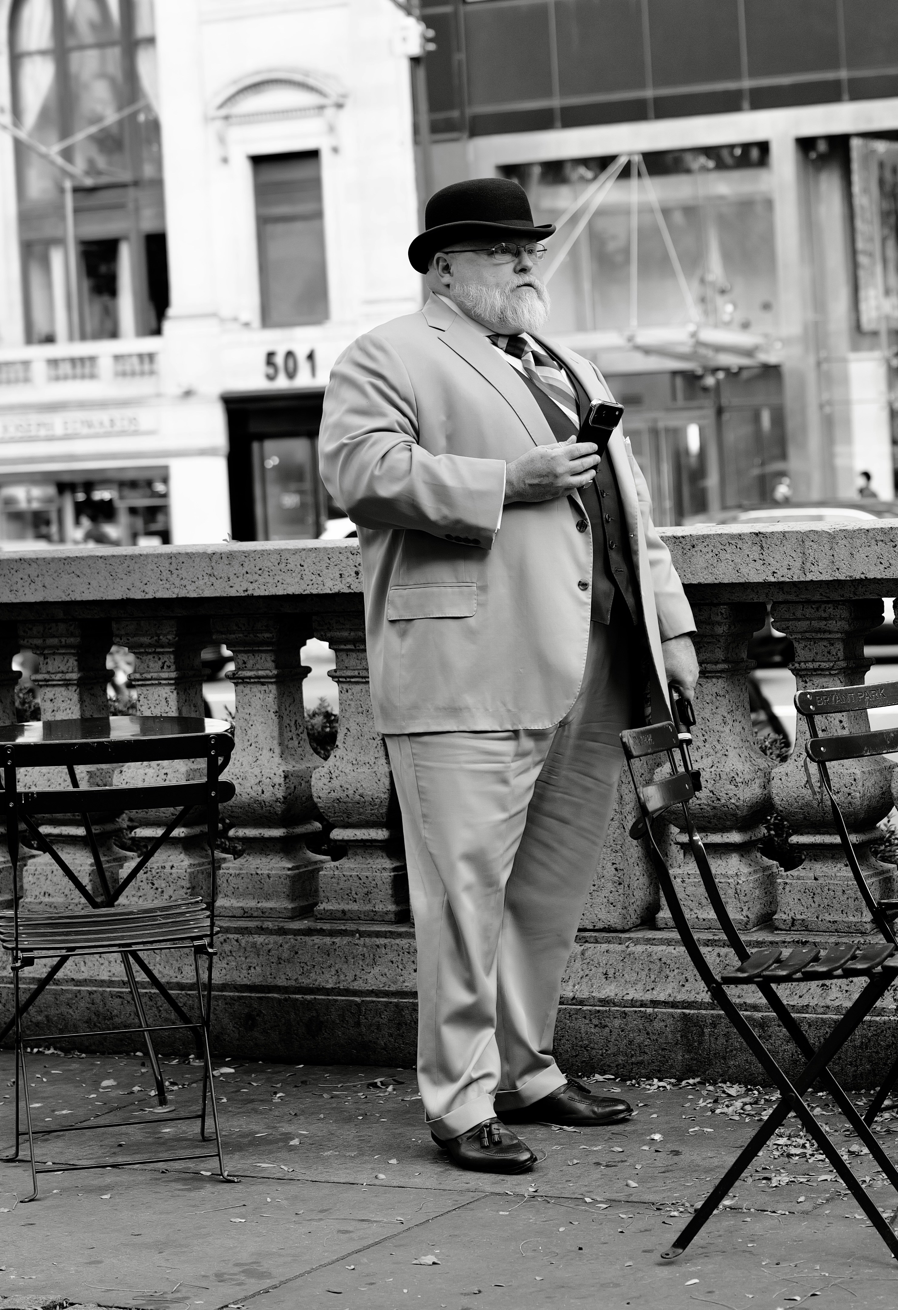 A man with a white beard in a suit and bowler hat holding a smartphone