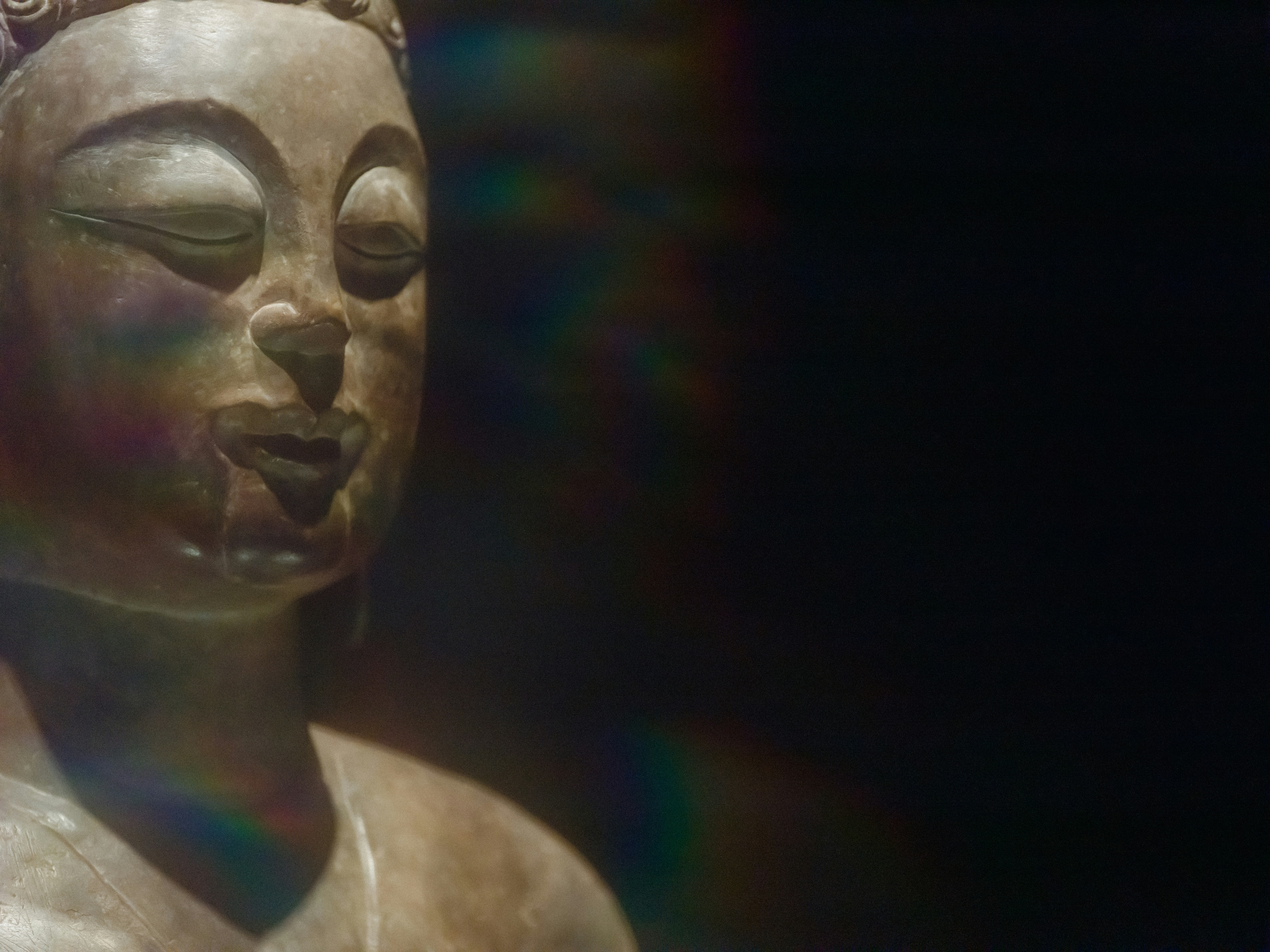 A stone Buddha statue with closed eyes illuminated by a soft rainbow light flare