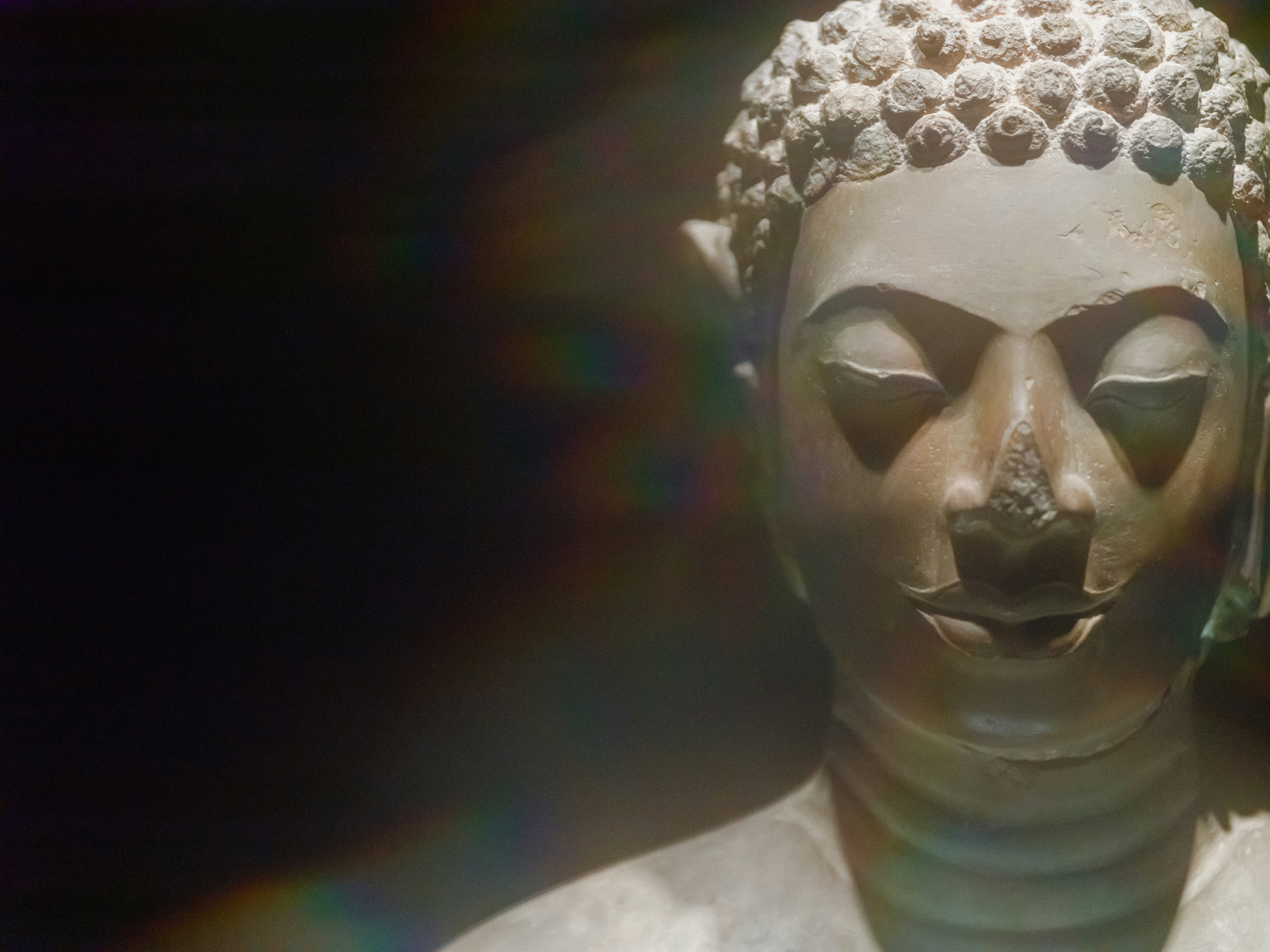 A stone Buddha statue head with closed eyes against a dark background with lens flare
