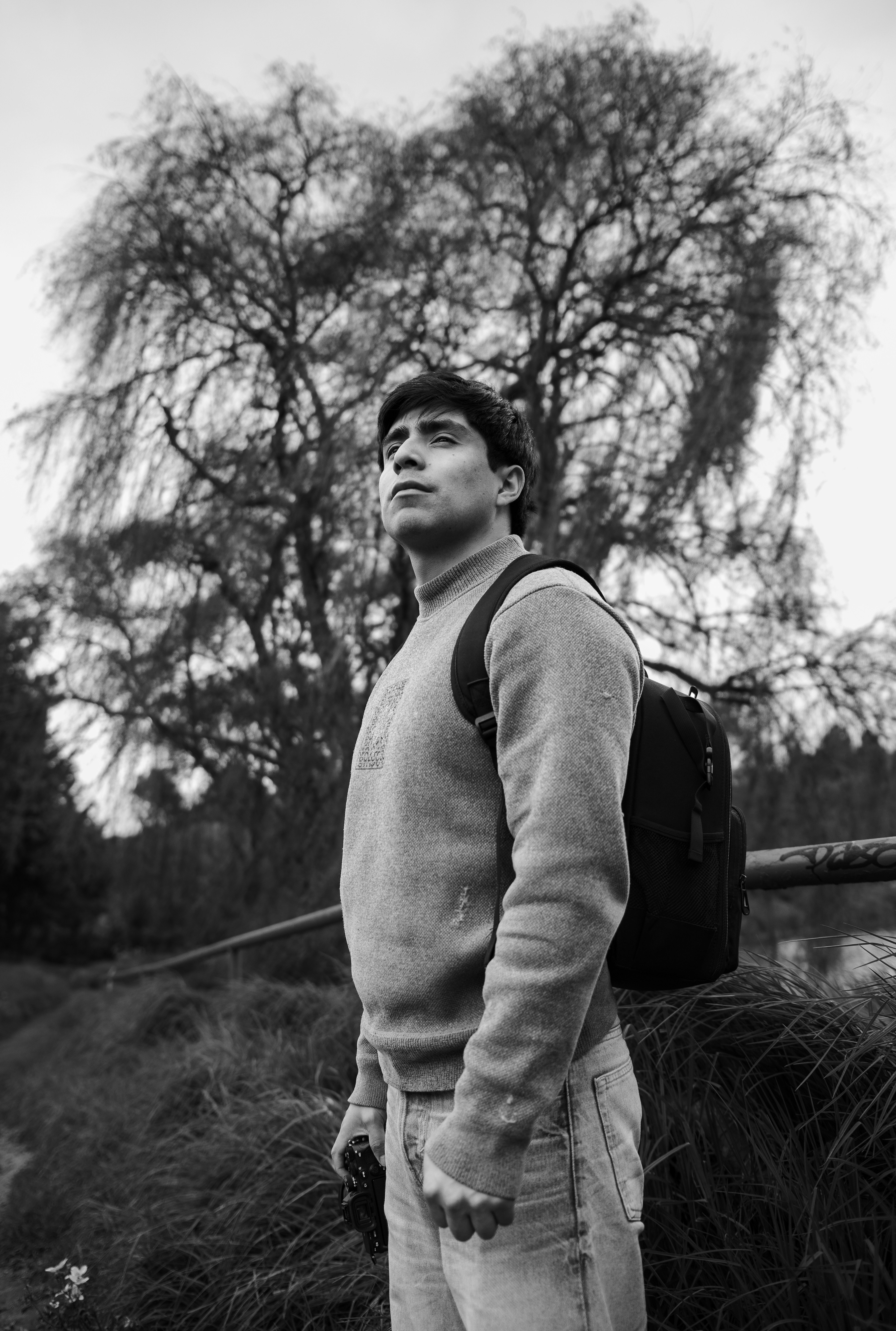 A man in a sweater and backpack holding a camera in a park