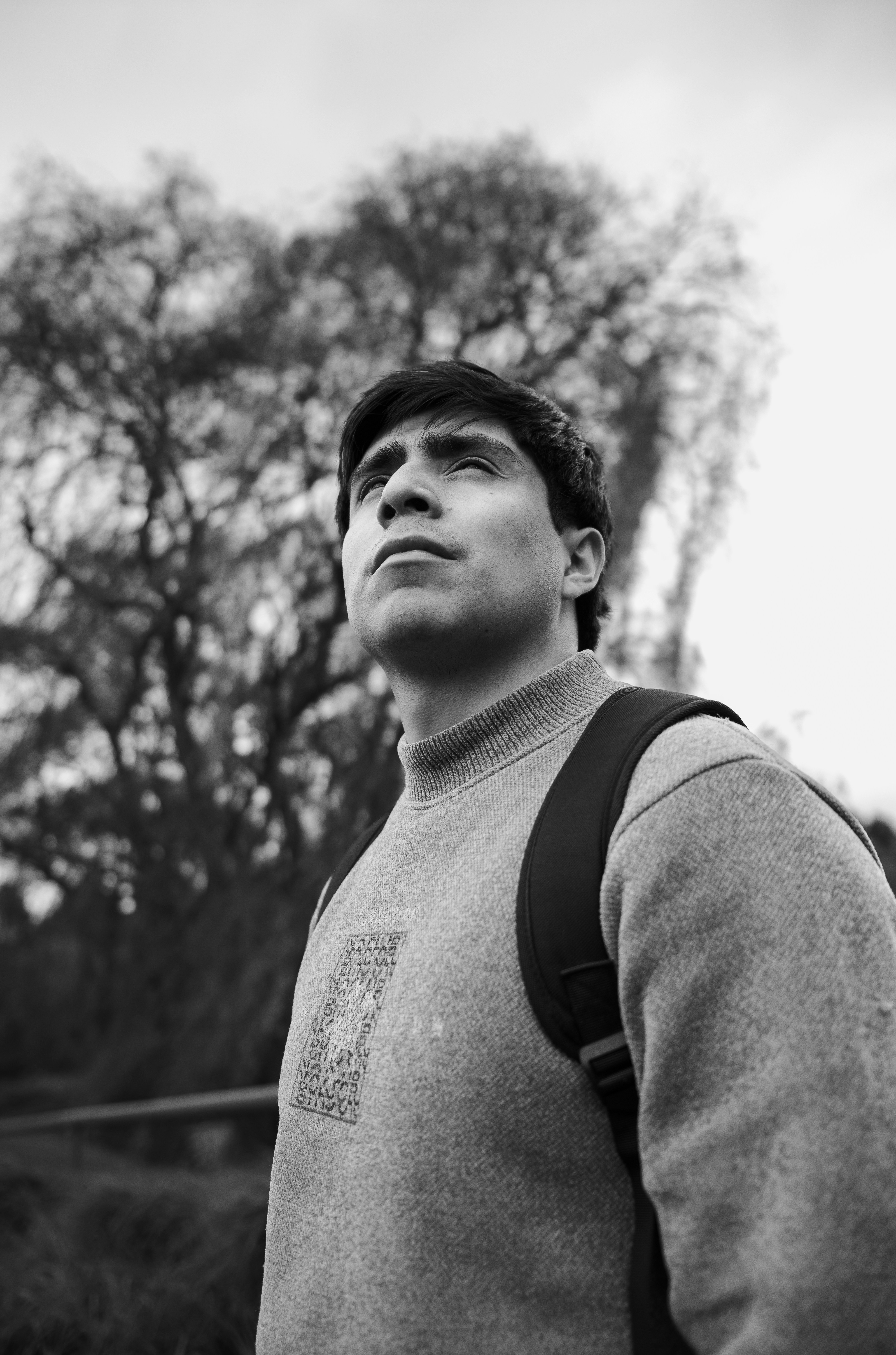 A young man in a grey sweater and backpack looking upward against blurred trees