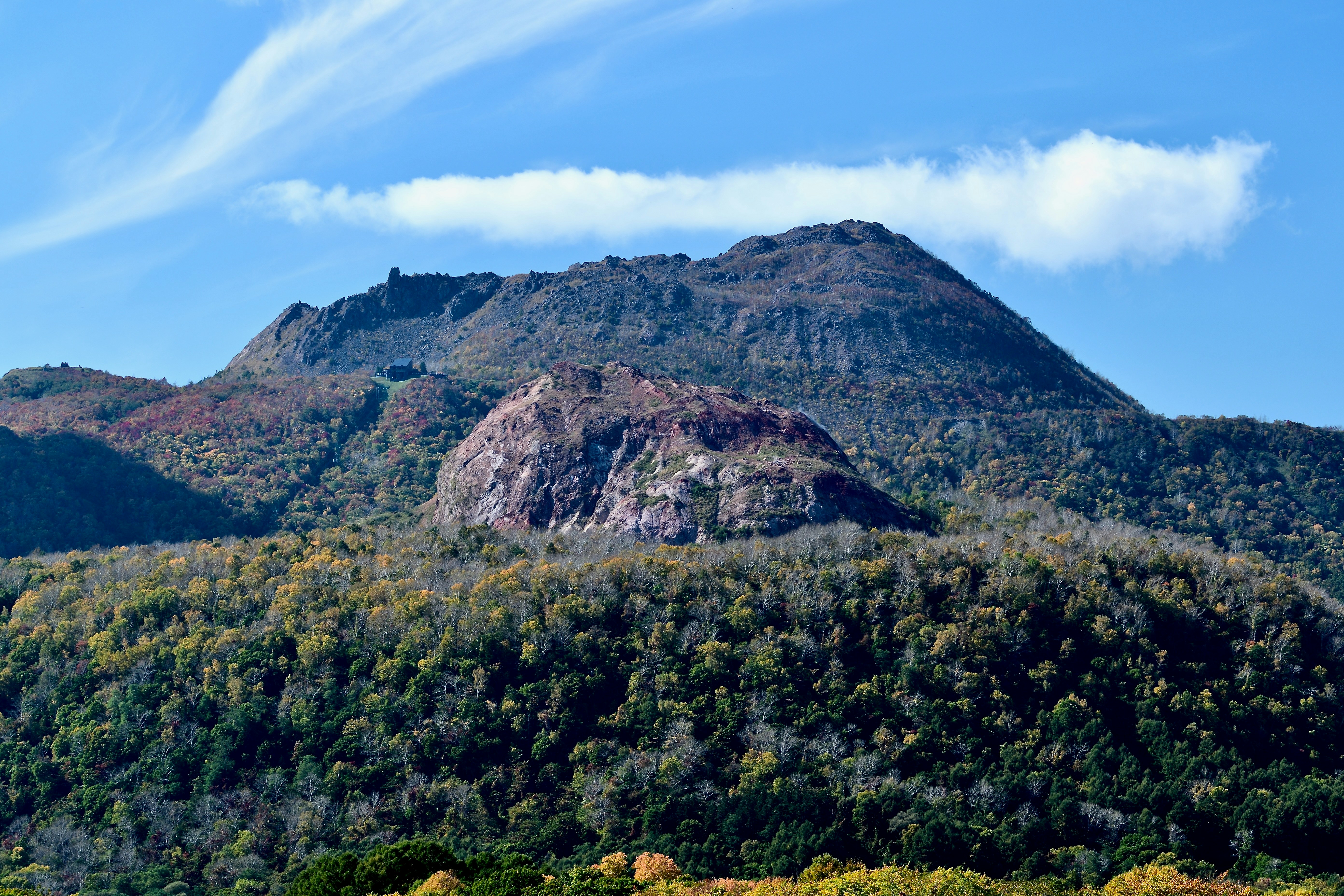 A rocky mountain peak rising above a dense forest with autumn foliage under a blue sky