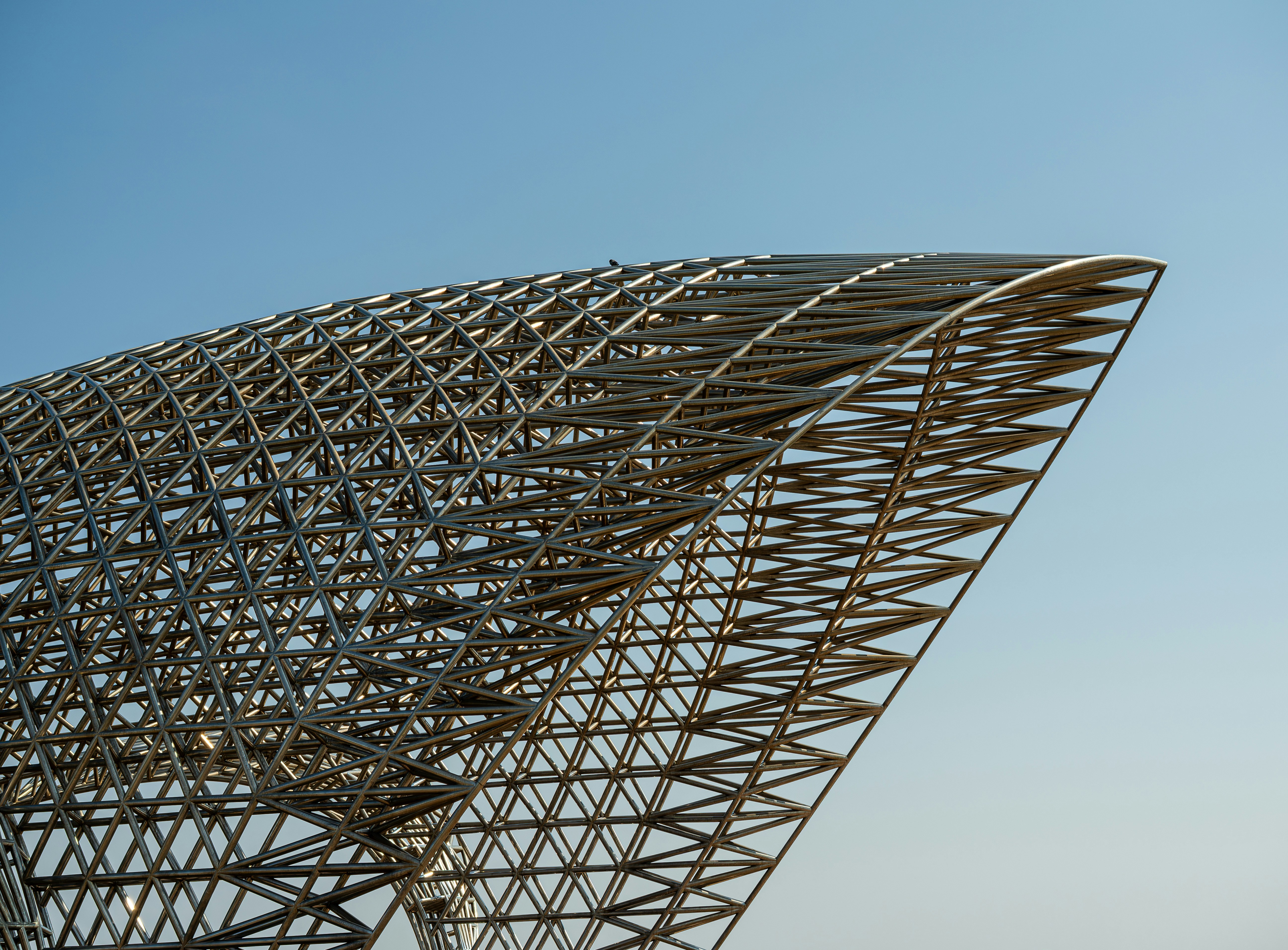 A complex metal lattice structure forming a curved geometric shape against a clear blue sky