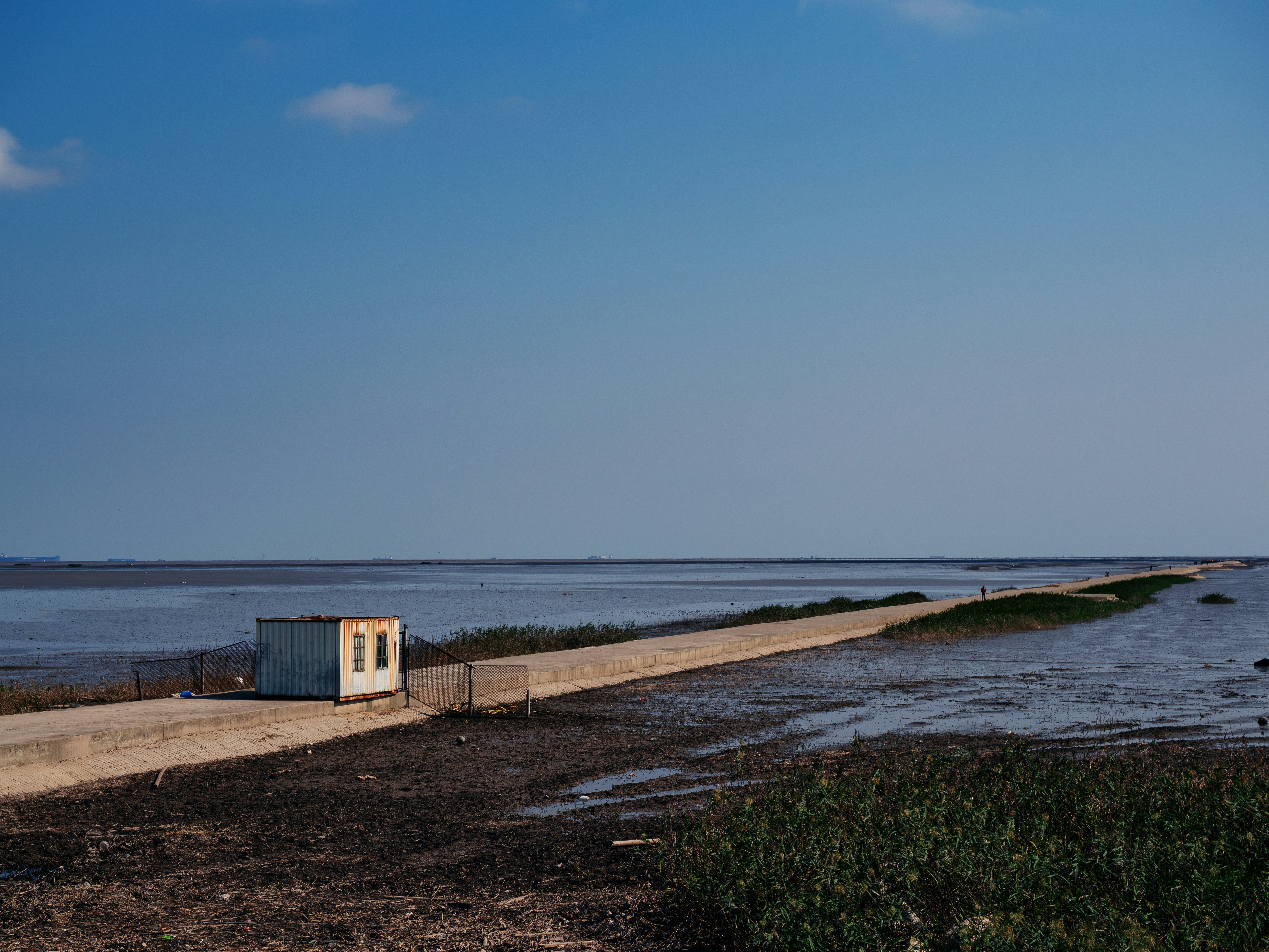 A small weathered container cabin on a long concrete causeway stretching into a vast tidal flat