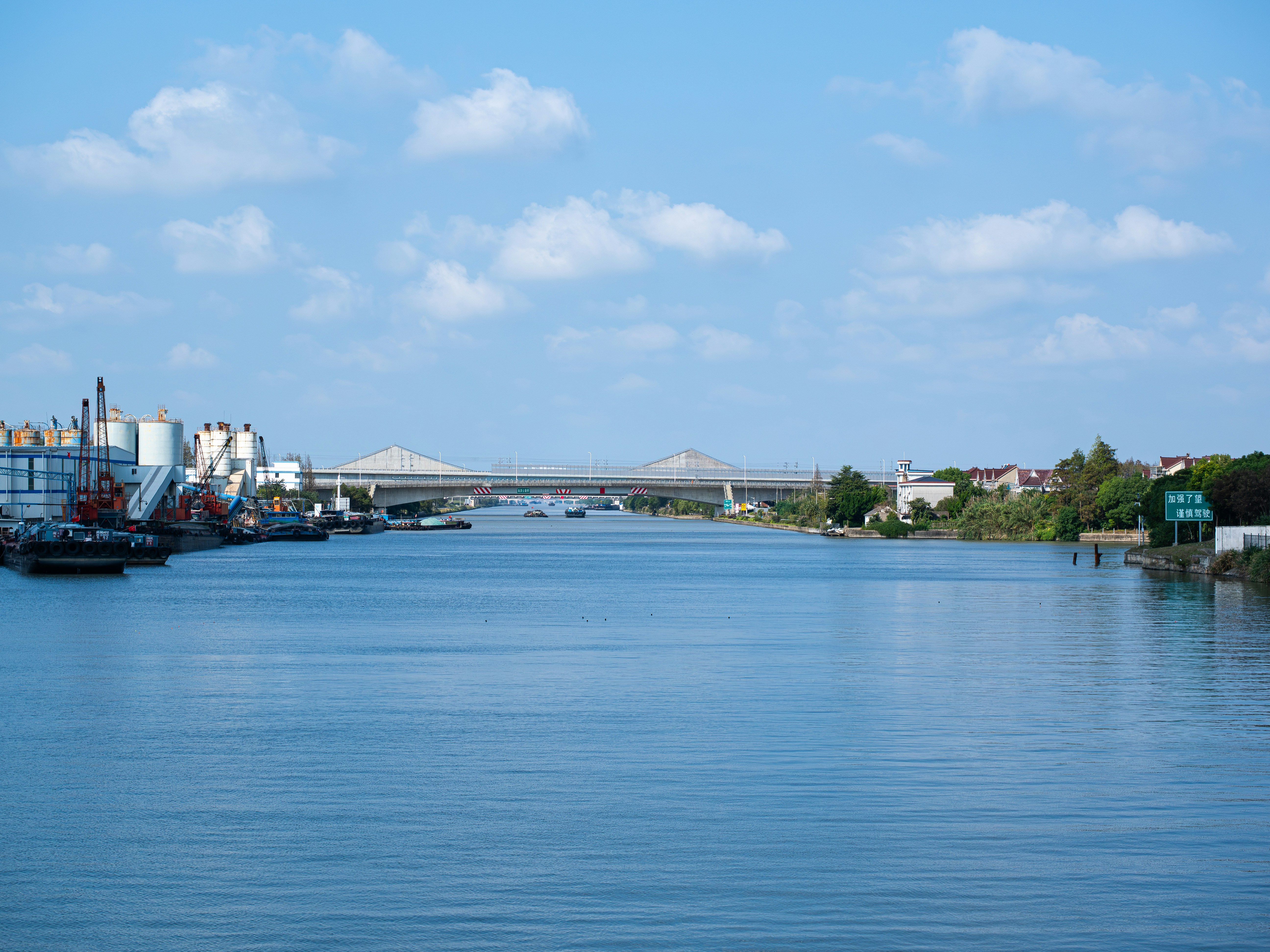 A wide industrial canal leading toward a concrete bridge under a partly cloudy sky