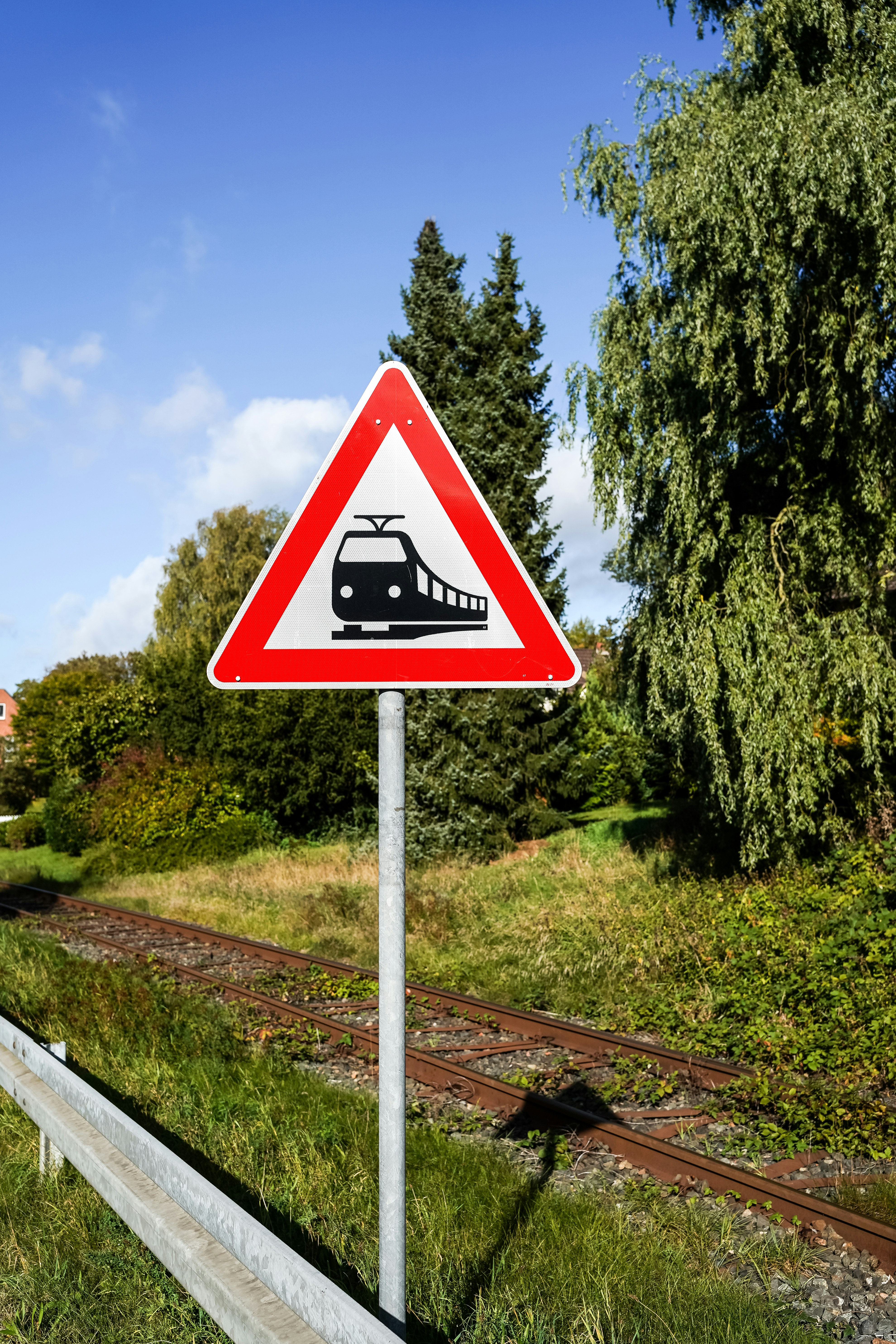 A triangular tram warning sign on a metal pole beside railway tracks and green trees